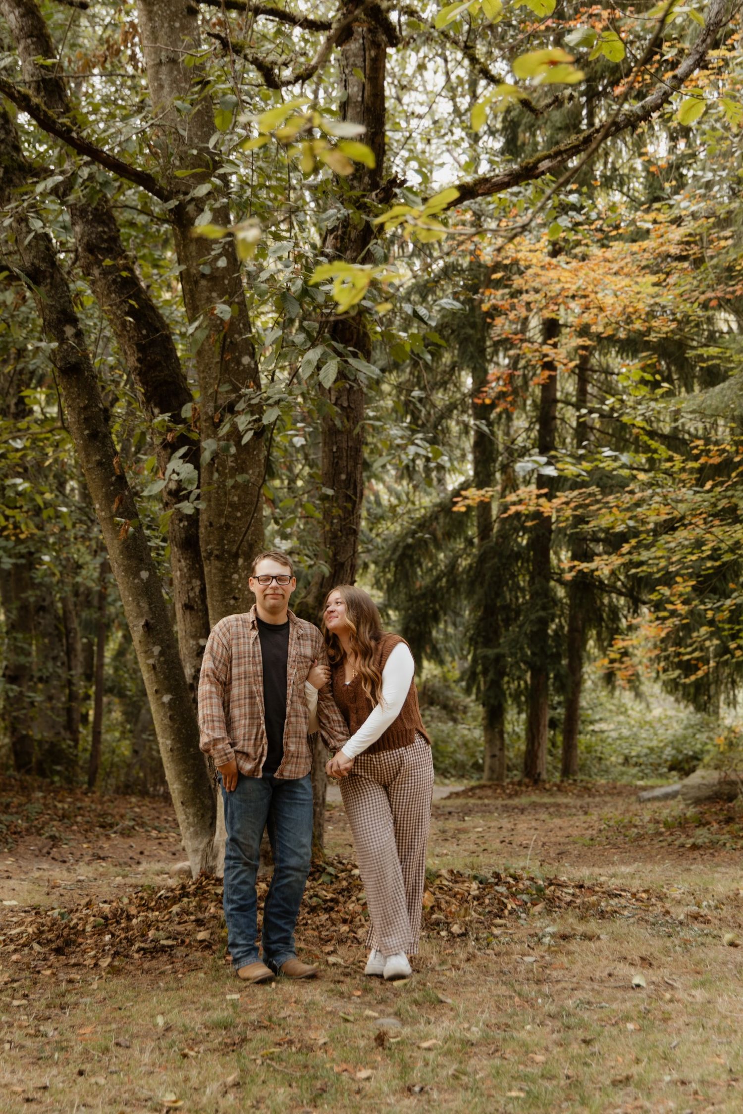 A couple stands together in an autumn forest setting with fallen leaves and mossy trees surrounding them.