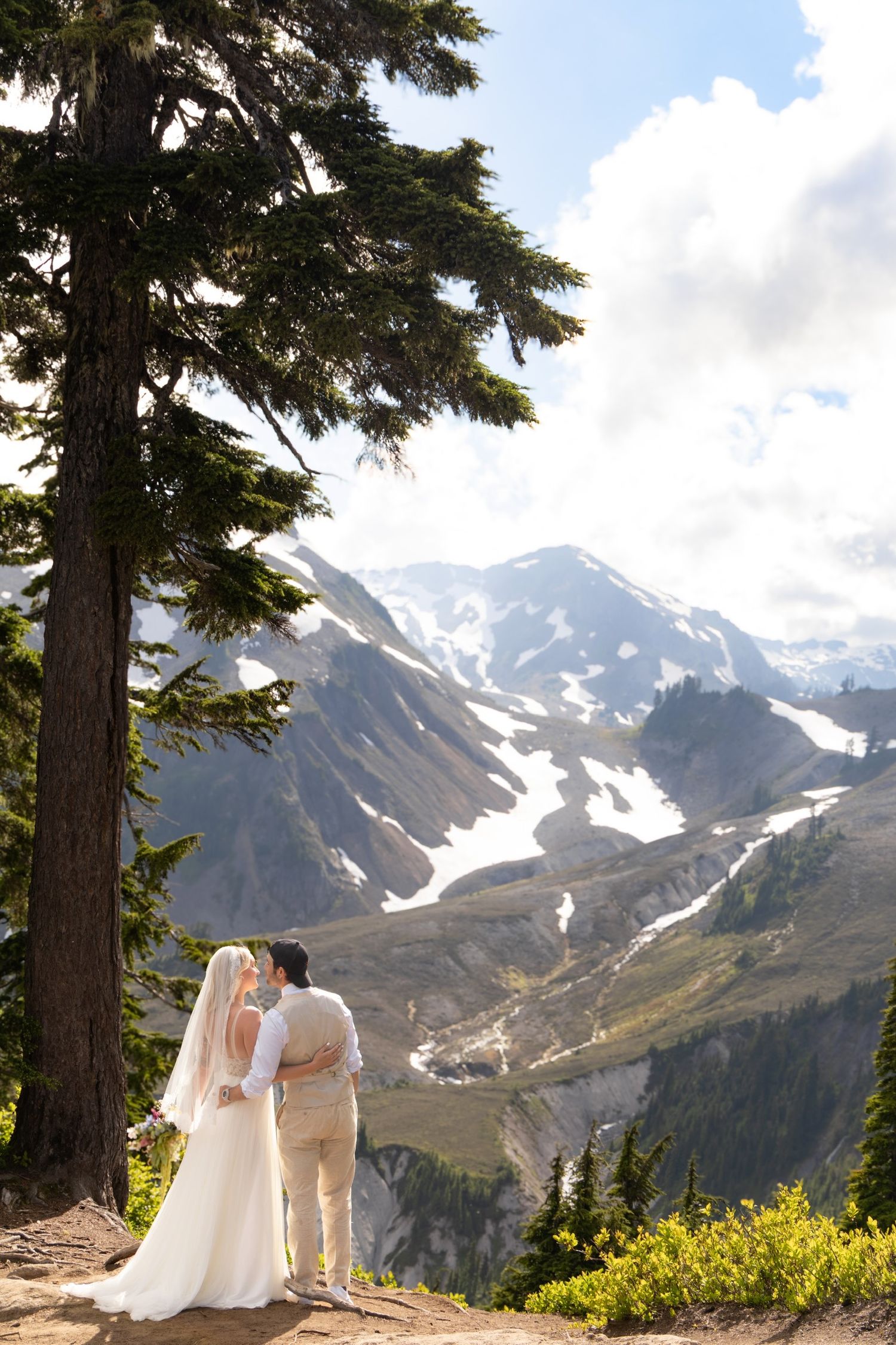 A couple stands at a scenic mountain viewpoint overlooking a snow-capped glacier valley in a dramatic alpine landscape.
