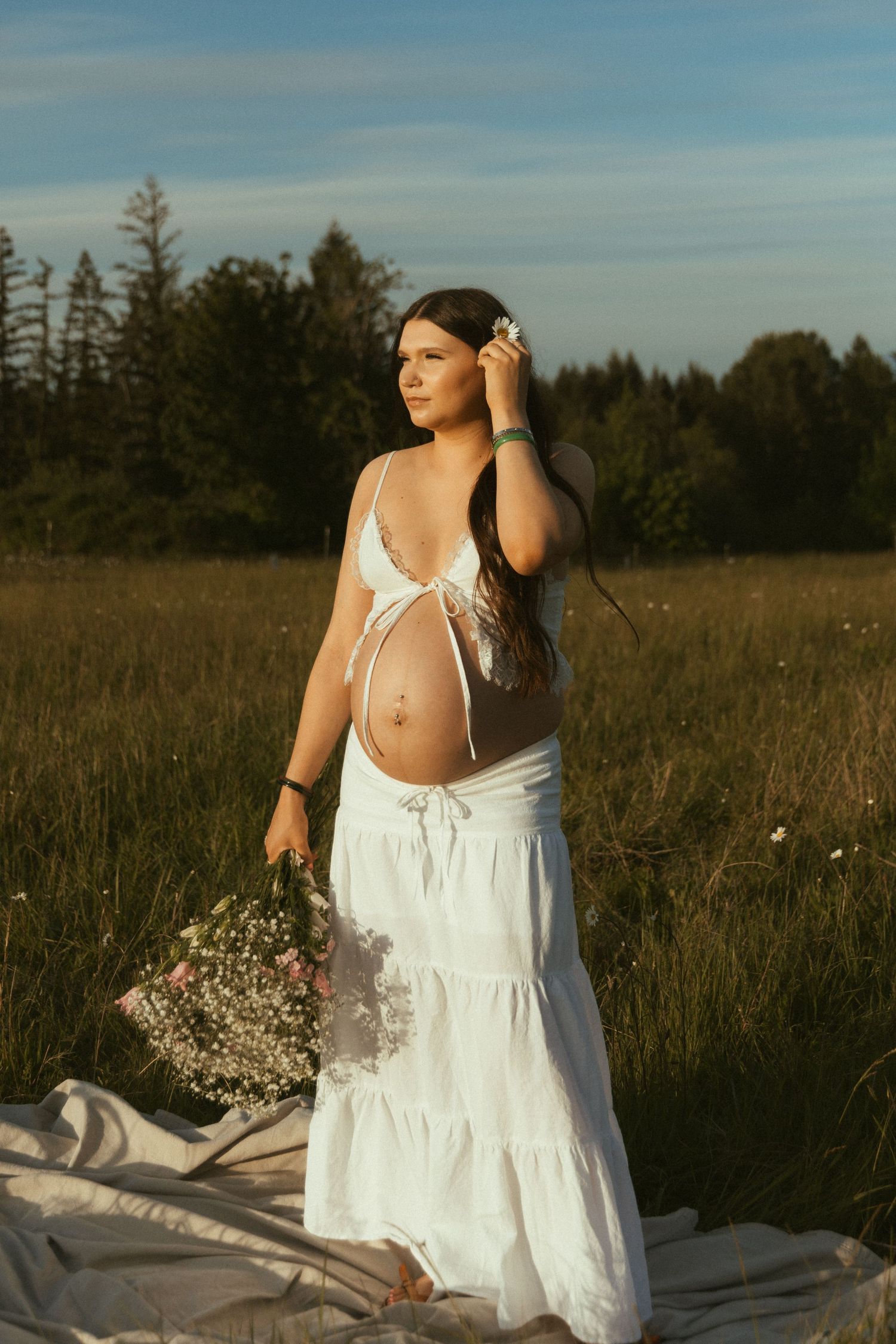 Pregnant woman in white crop top and maxi skirt holding wildflowers in a sunlit field at golden hour.