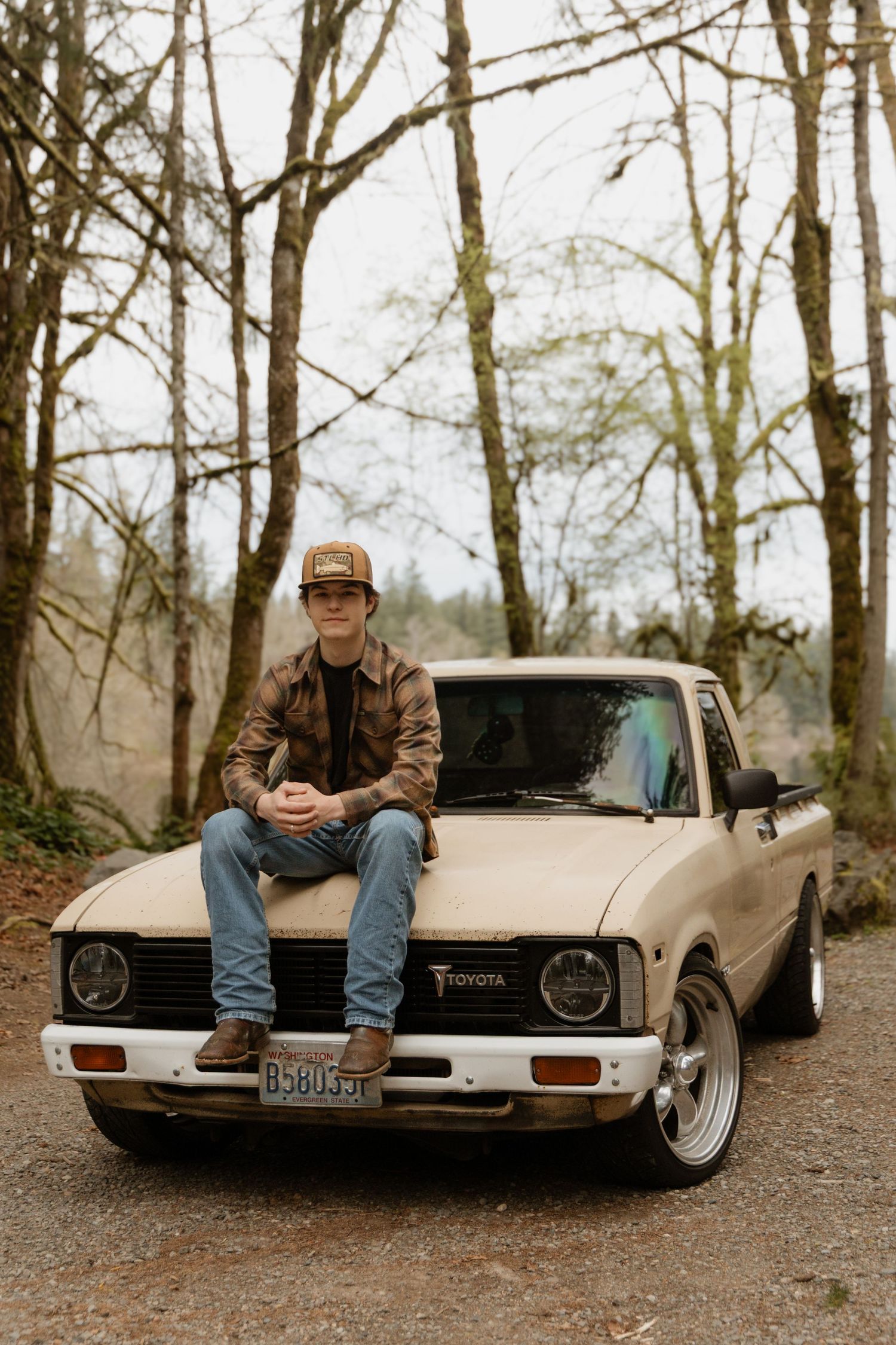 Young man in cap and camo jacket sitting on hood of vintage Toyota pickup truck on a forest road.
