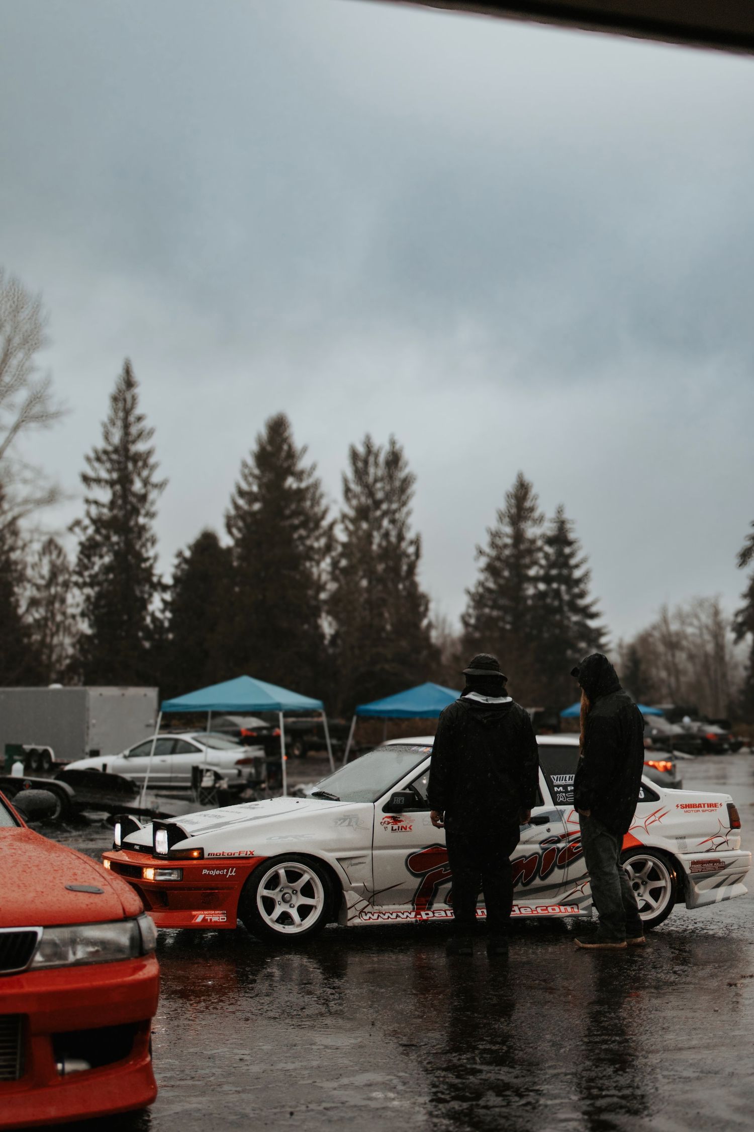 Car enthusiasts gather at a rainy meetup to admire custom vehicles and modified cars in an overcast parking lot.