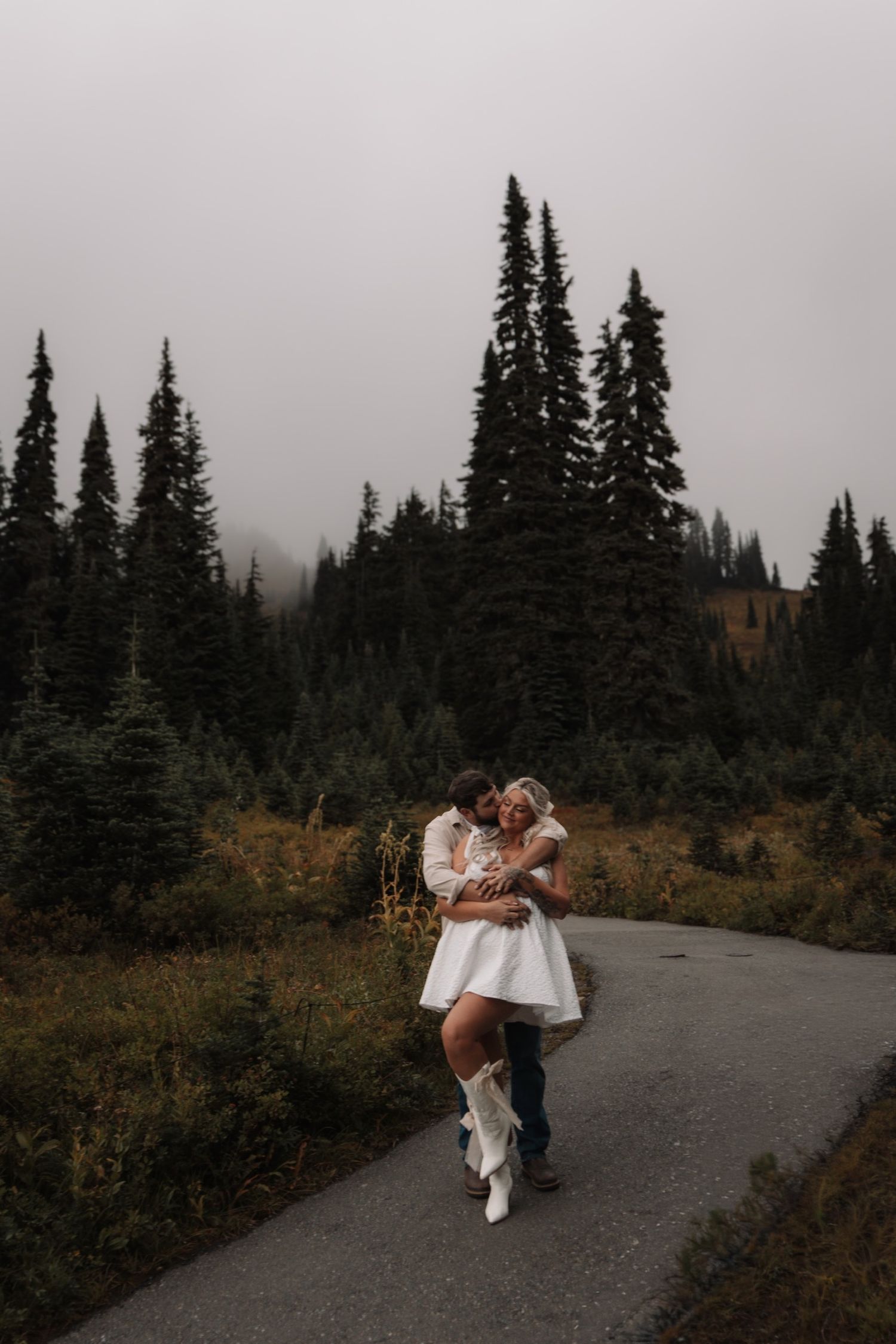 A couple sharing a playful moment on a misty mountain path surrounded by pine trees.