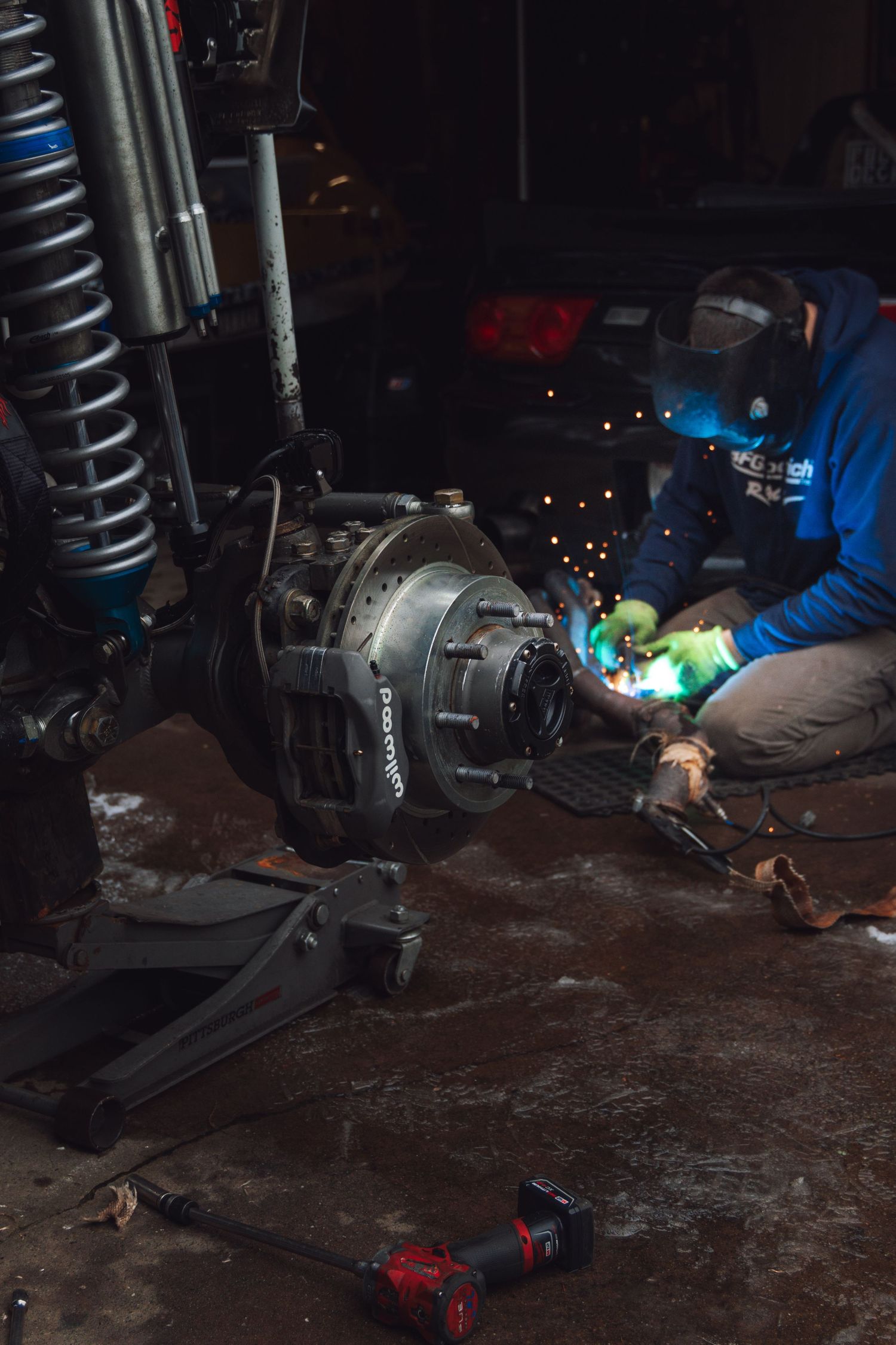 Mechanic welding automotive parts in a dark garage workshop with sparks flying.