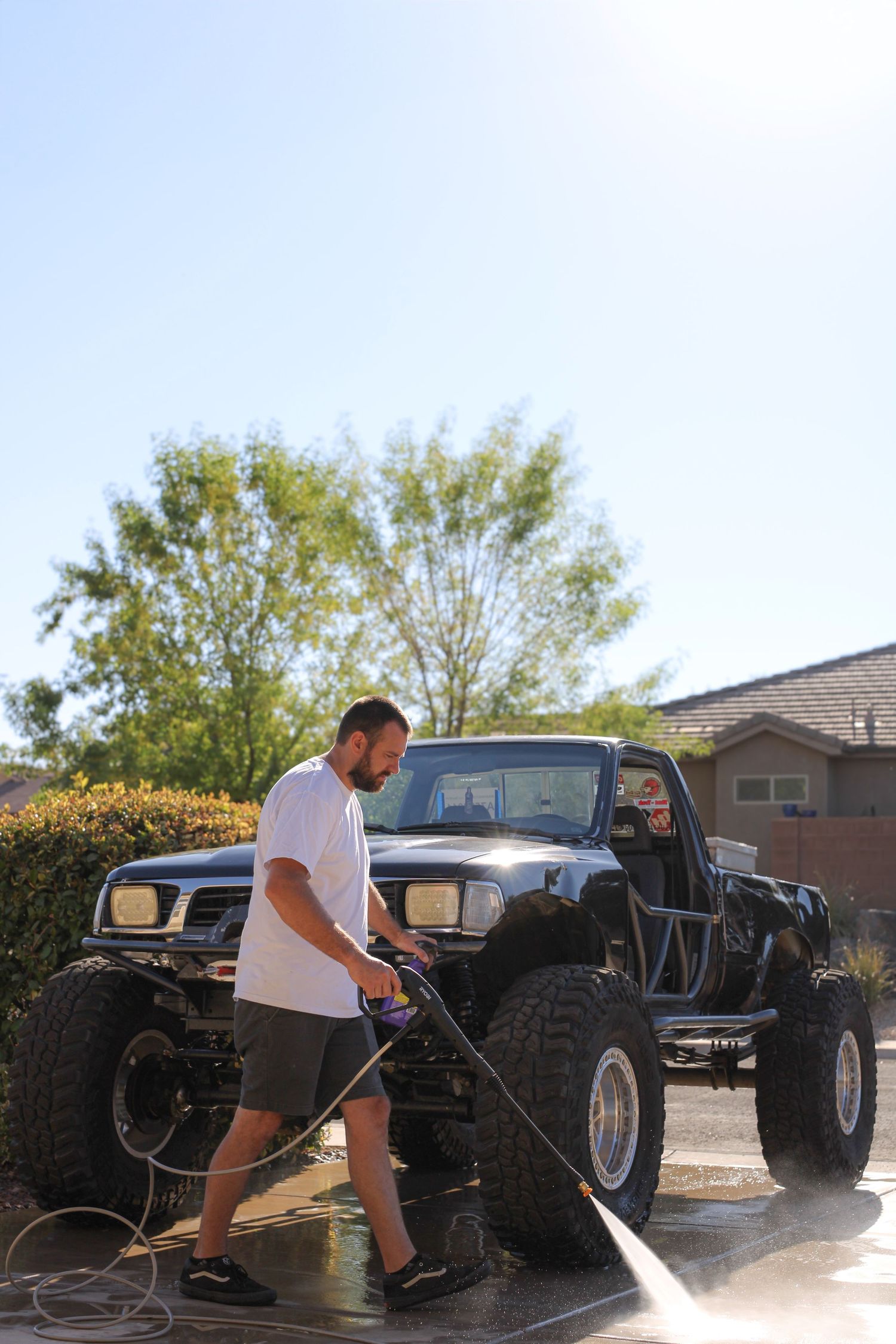 Someone washing a lifted black pickup truck with a pressure washer in a driveway on a sunny day.
