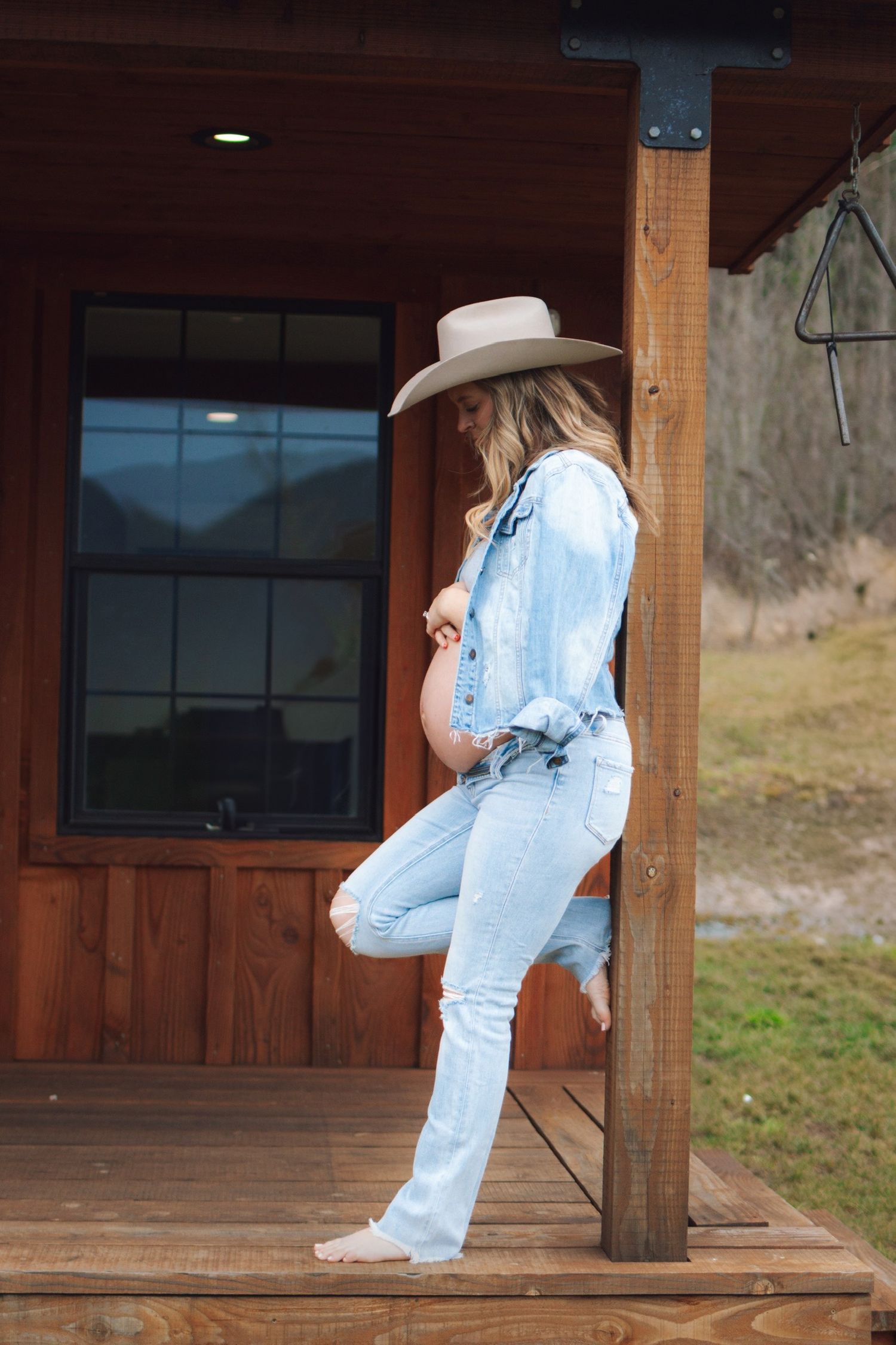 Pregnant woman in denim and cowboy hat leaning against rustic wooden post.