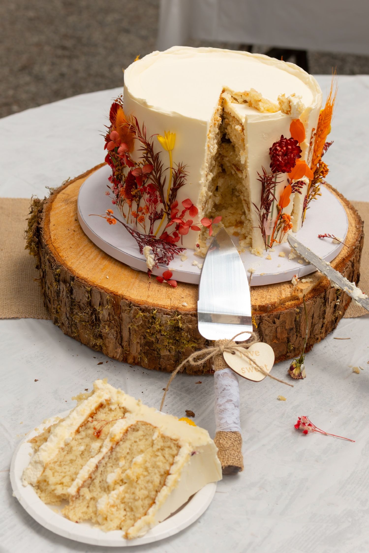 A rustic wedding cake decorated with dried flowers on a wood slice serving board.