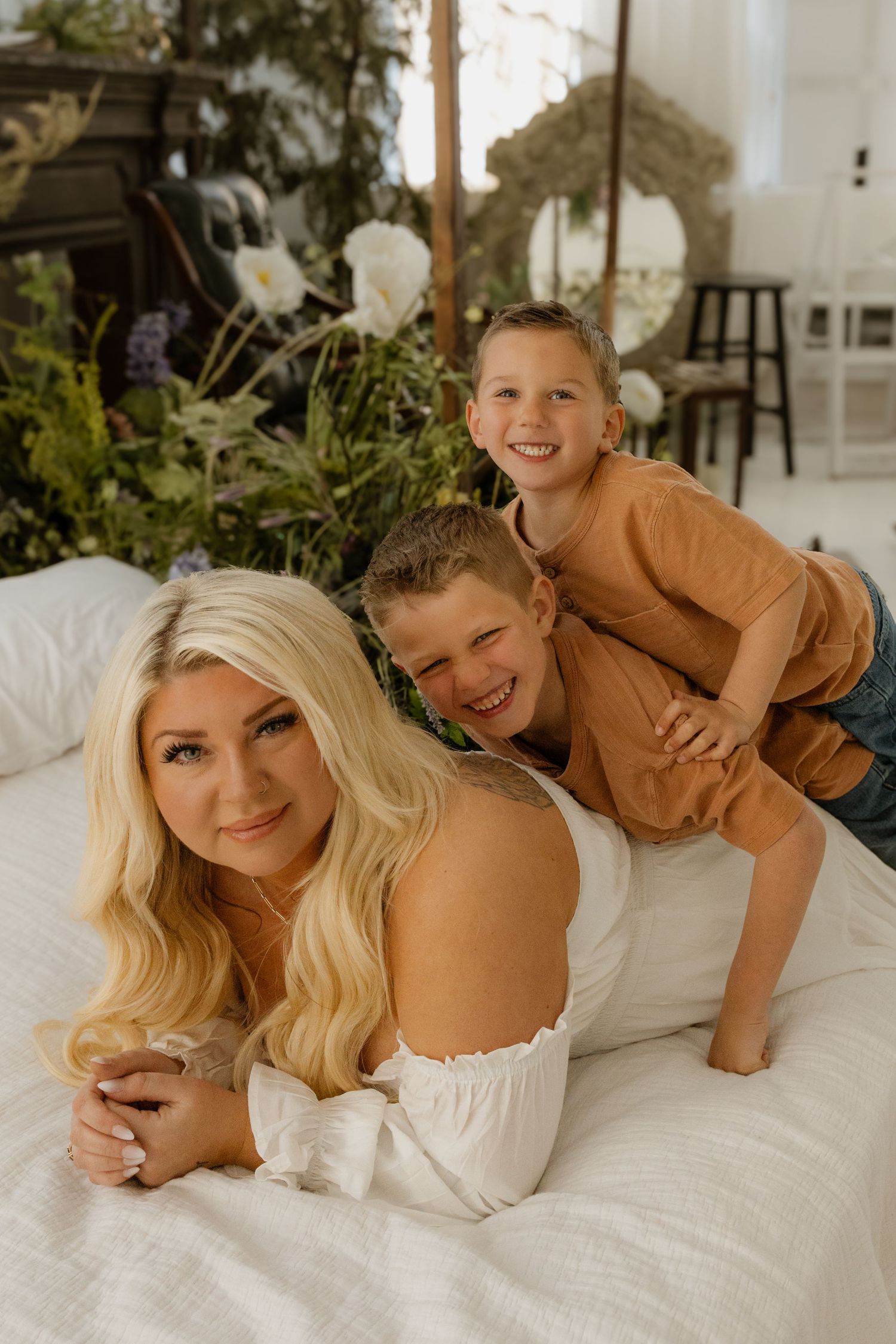 Blonde woman in white dress lying on bed smiling with two young boys laughing behind her in a bright studio setting.