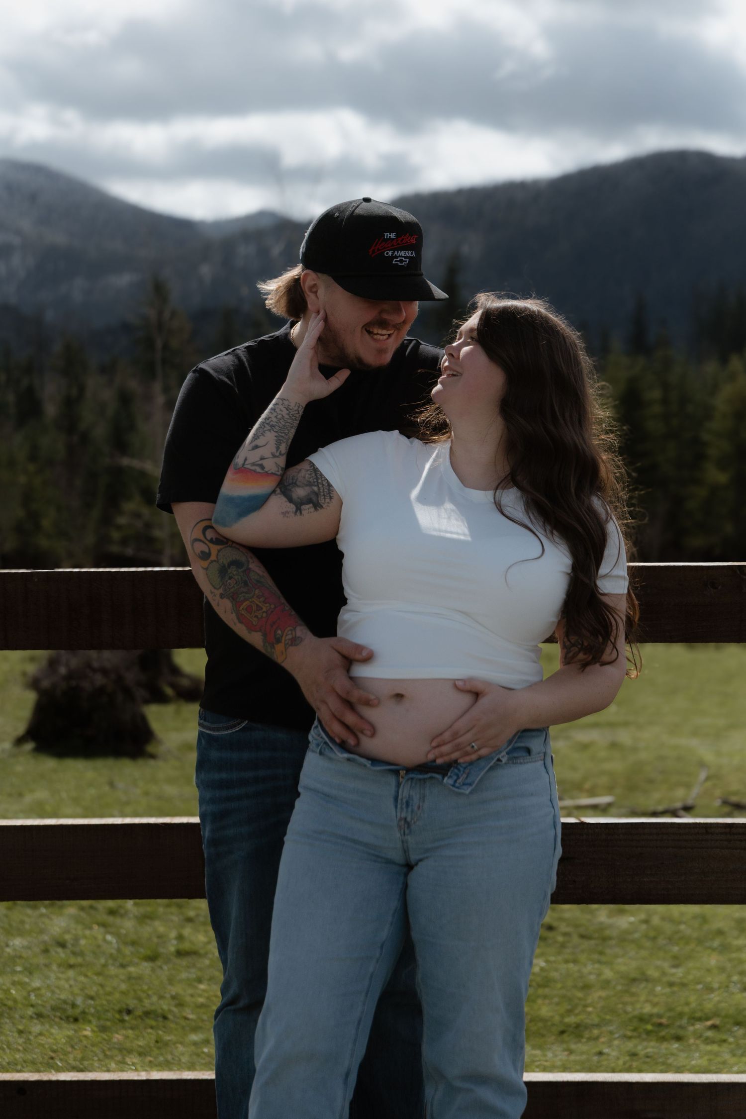 A romantic couple shares tender moments in a mountain meadow with evergreen trees and cloudy skies in the background.