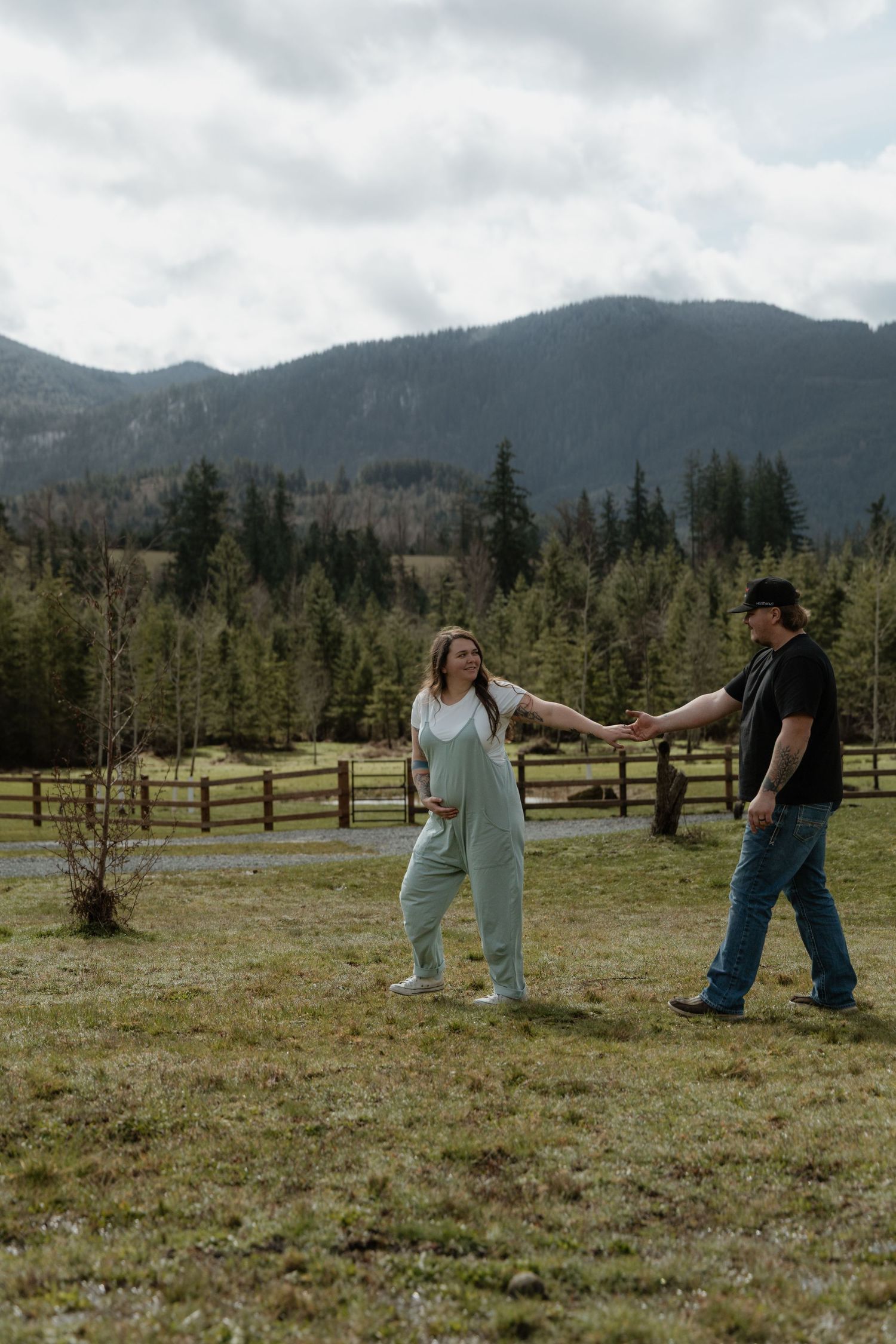 A couple holds hands and spins playfully in a mountain meadow with evergreen trees and misty peaks in the background.