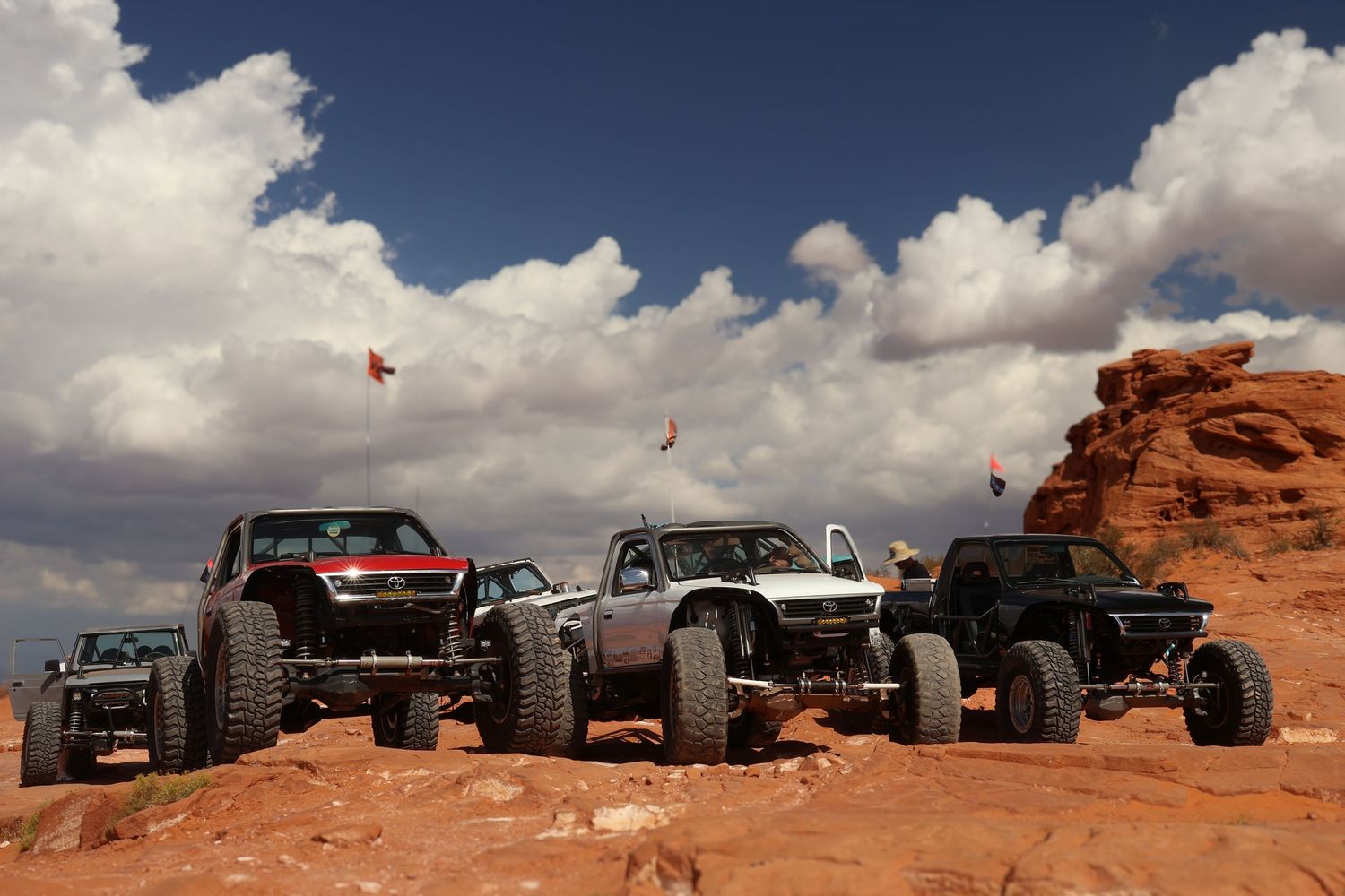 Off-road vehicles traverse rugged red rock terrain under dramatic cloudy skies in a desert landscape.