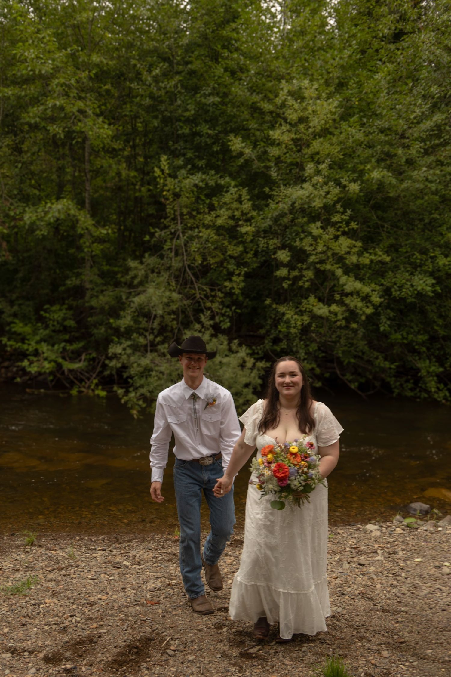 A couple in wedding attire walks along a riverbank surrounded by lush green trees on their special day.