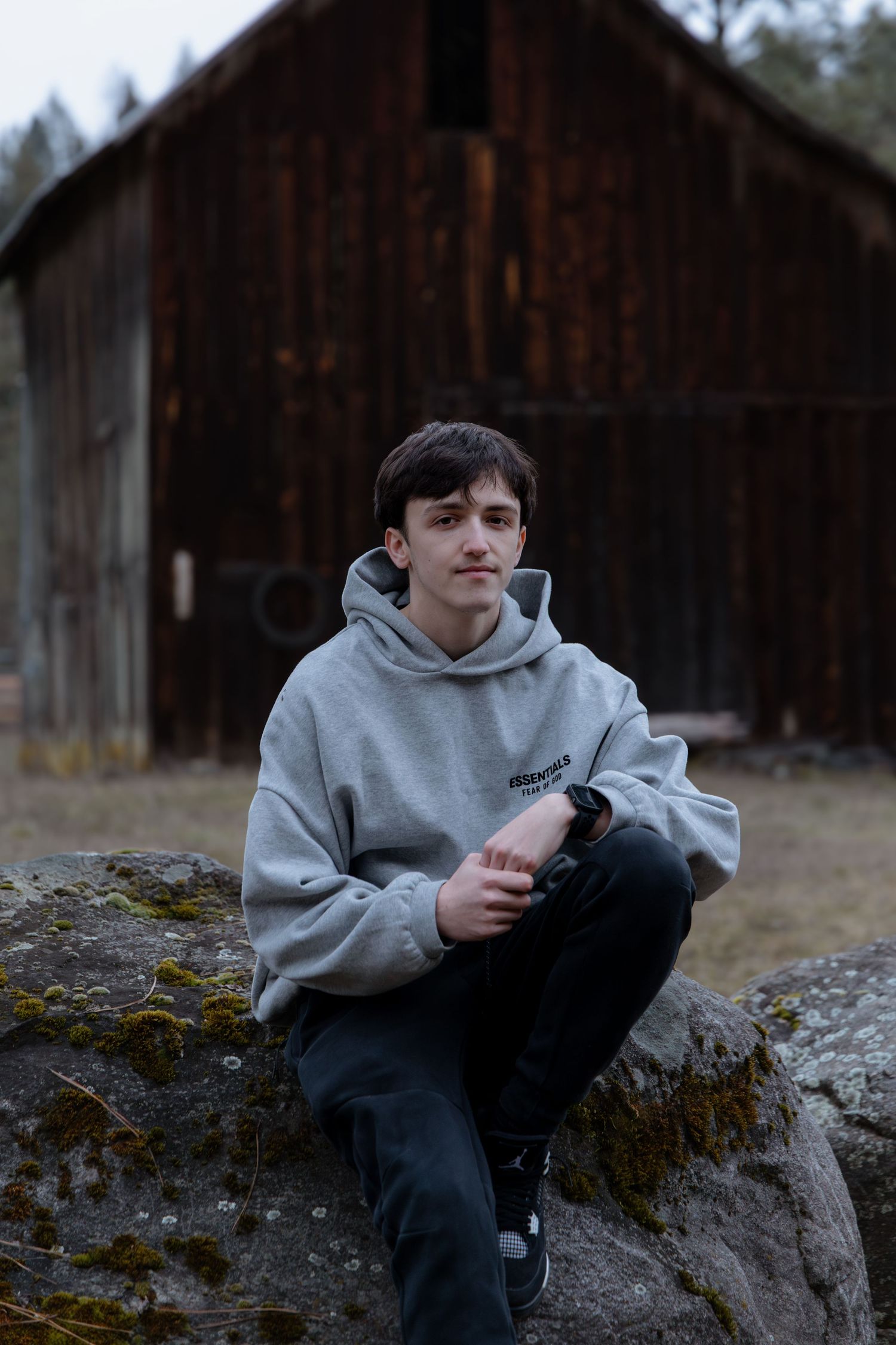 A person in a gray hoodie sits on a moss-covered rock in a wooded area with rustic buildings in the background.