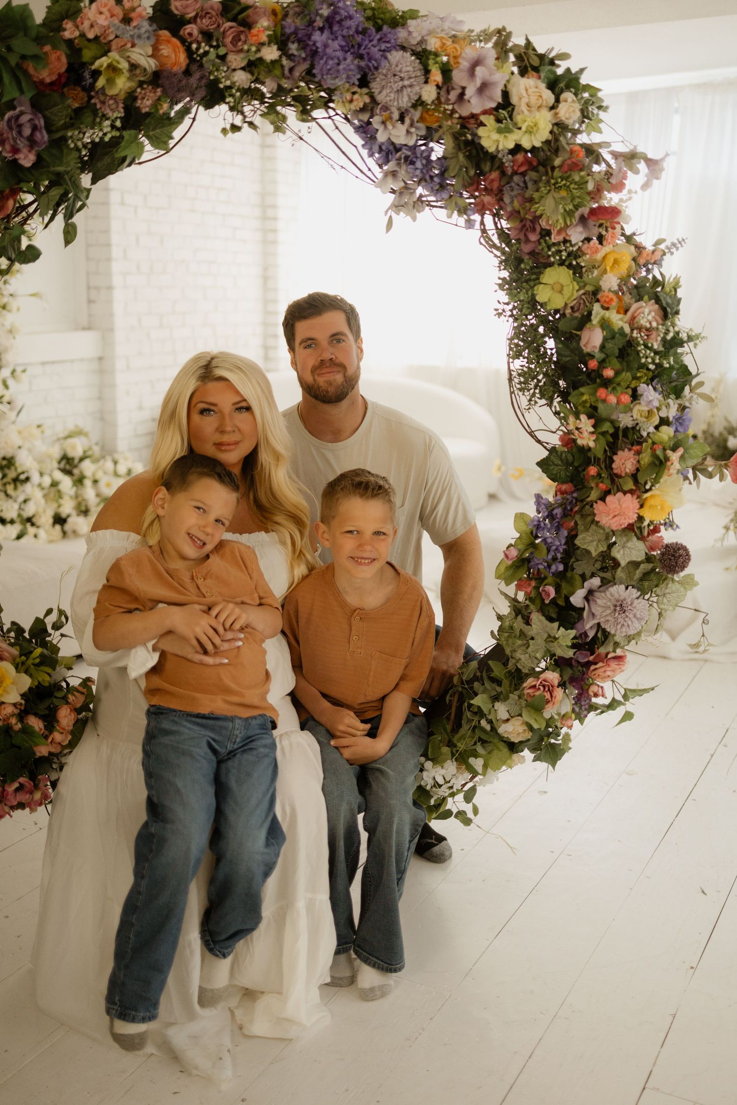 Happy family of four posing under a colorful floral arch in a bright white studio setting.