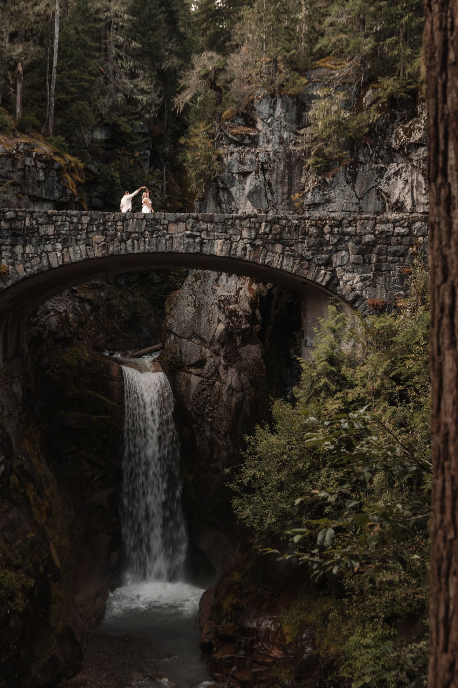A stone bridge spans over a dramatic waterfall in a lush forest setting.