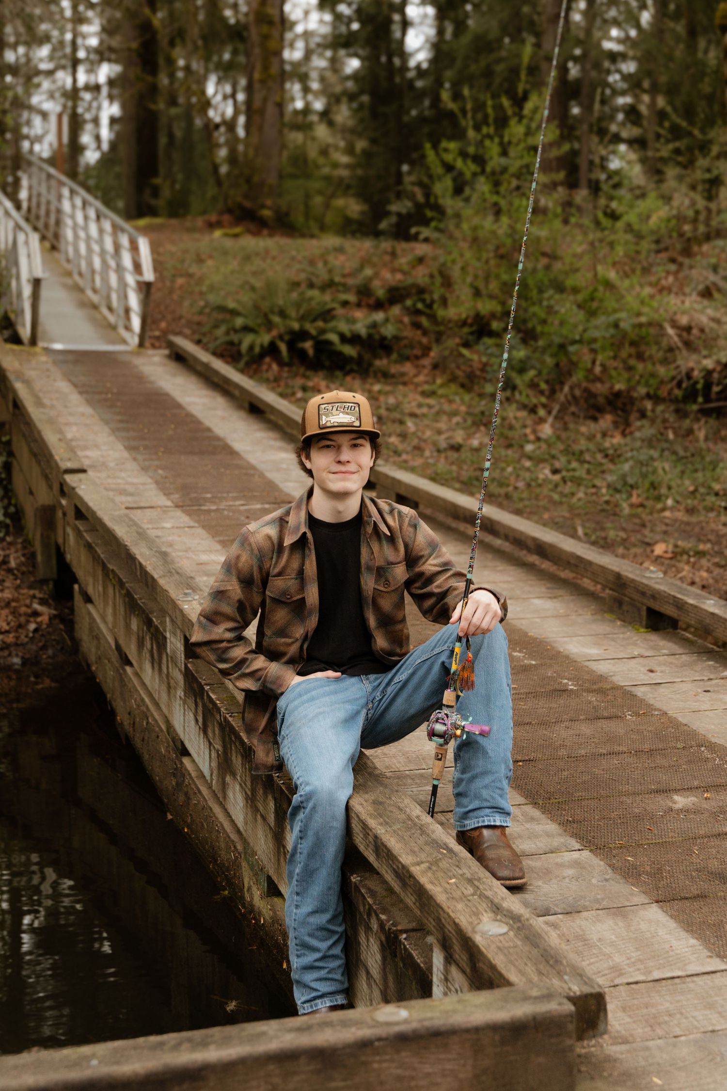 Young man in plaid shirt and trucker hat sitting on wooden bridge with fishing rod in wooded area.