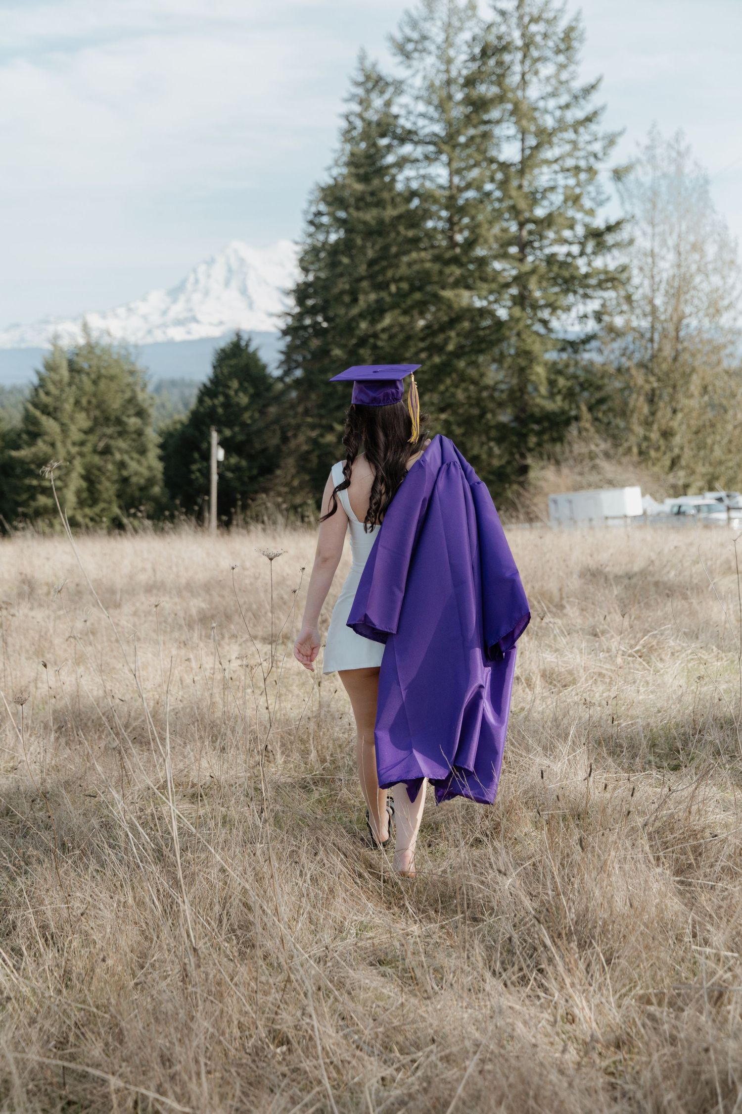 A person wearing a purple graduation gown walks through a dry grass field on a cloudy day.