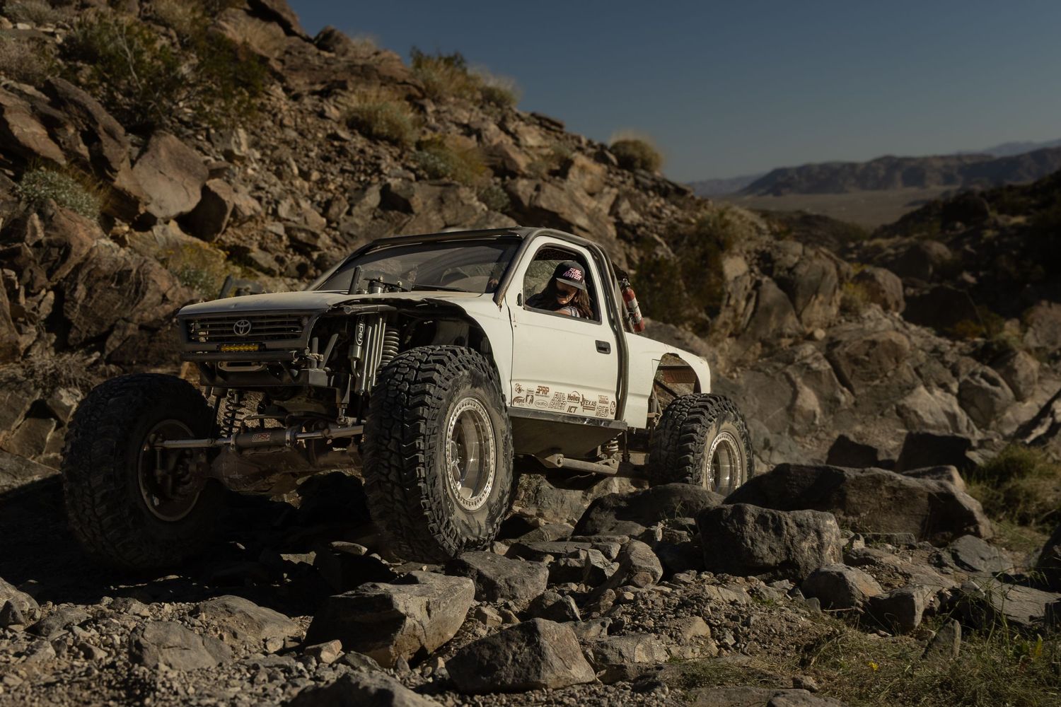 White off-road truck navigates rocky desert terrain with steep cliffs and boulders in the background.