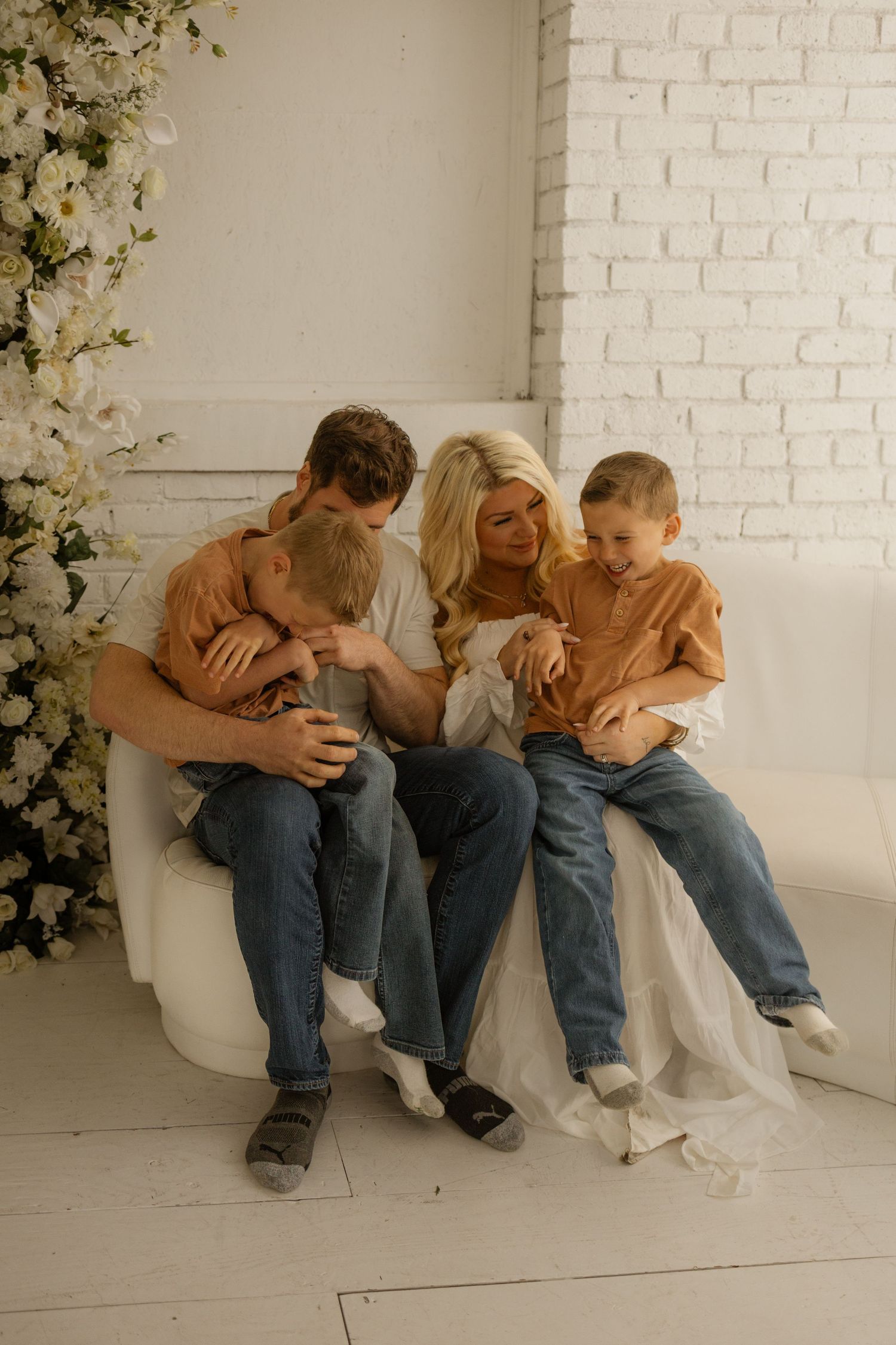 A happy family of four sits together on a white chair surrounded by white flowers, parents hugging two young boys in matching outfits.