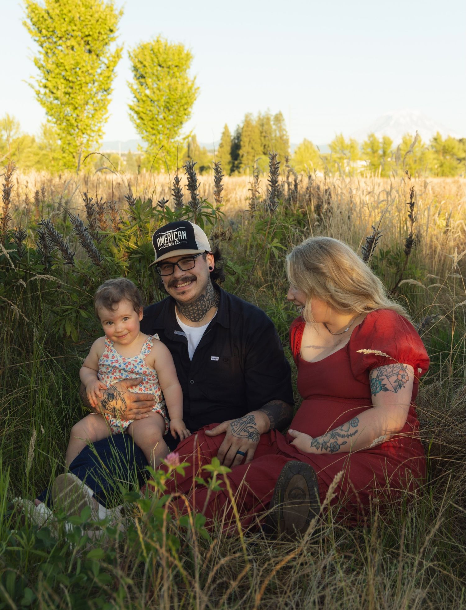 A family sits together in tall grass during golden hour, sharing tender moments in a natural outdoor setting.