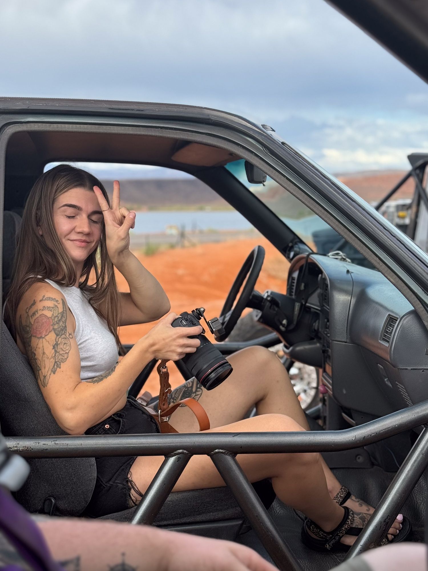 Person sitting in driver's seat of car taking photos with camera equipment in desert landscape setting.
