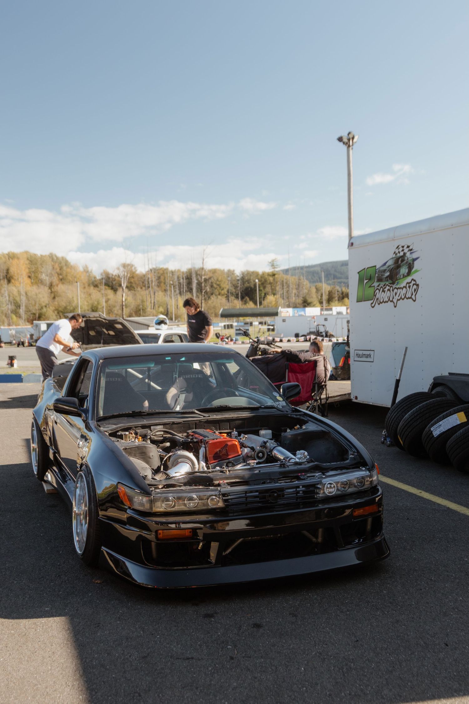 Multiple black sports cars displayed at an outdoor car meet with trailers and mountains visible in the background.