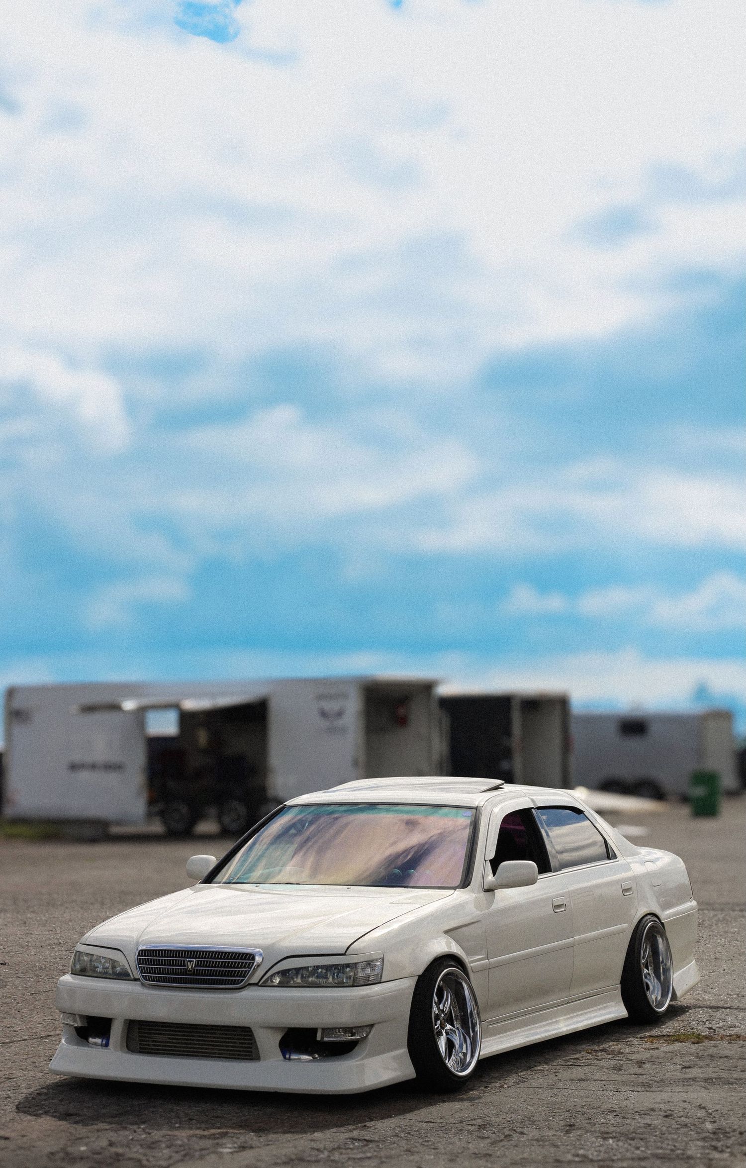 Four sequential views of a lowered white custom model car on a concrete surface against a cloudy blue sky background.