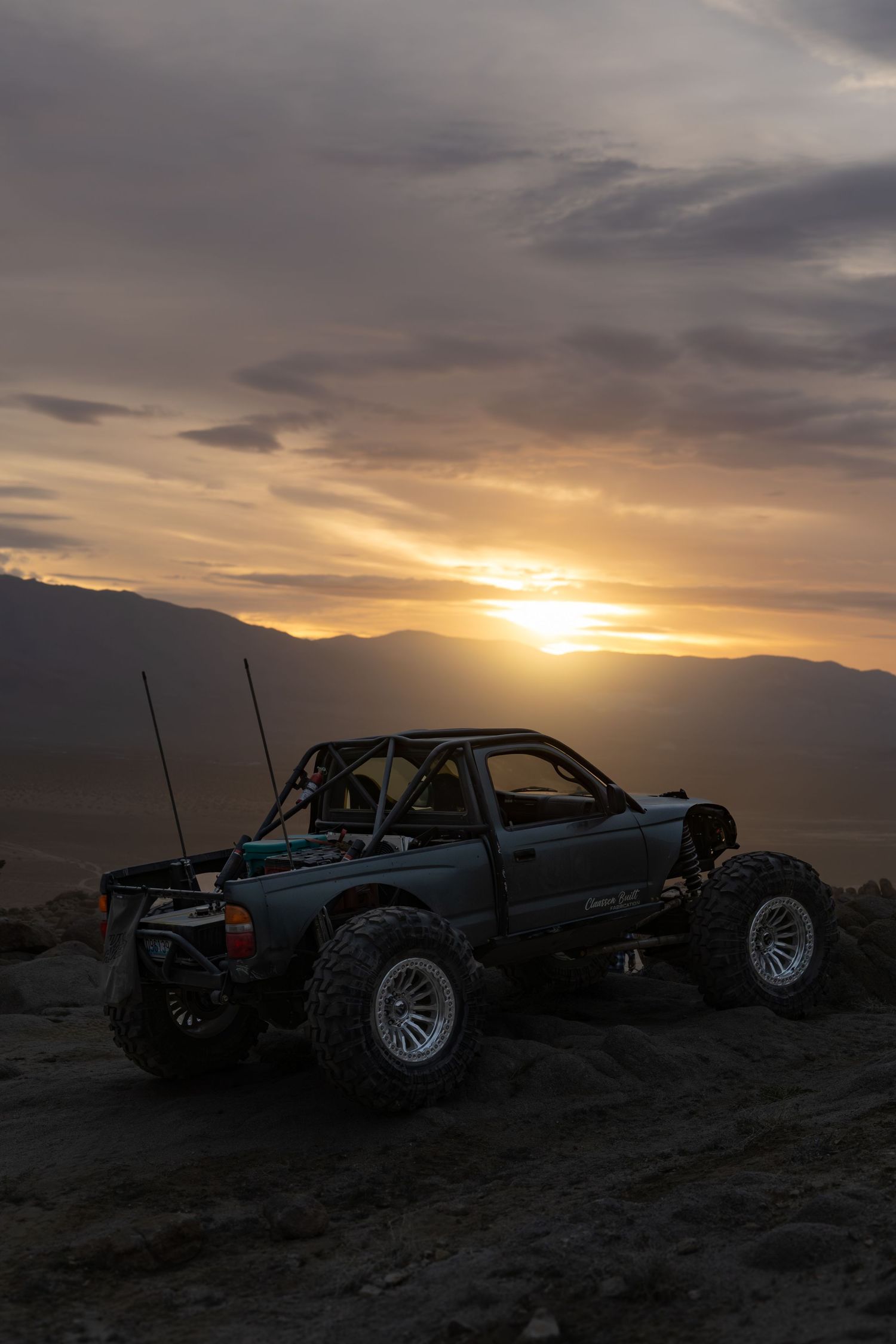 Silhouetted off-road vehicles parked on a hilltop against a dramatic sunset sky with mountains in the background.