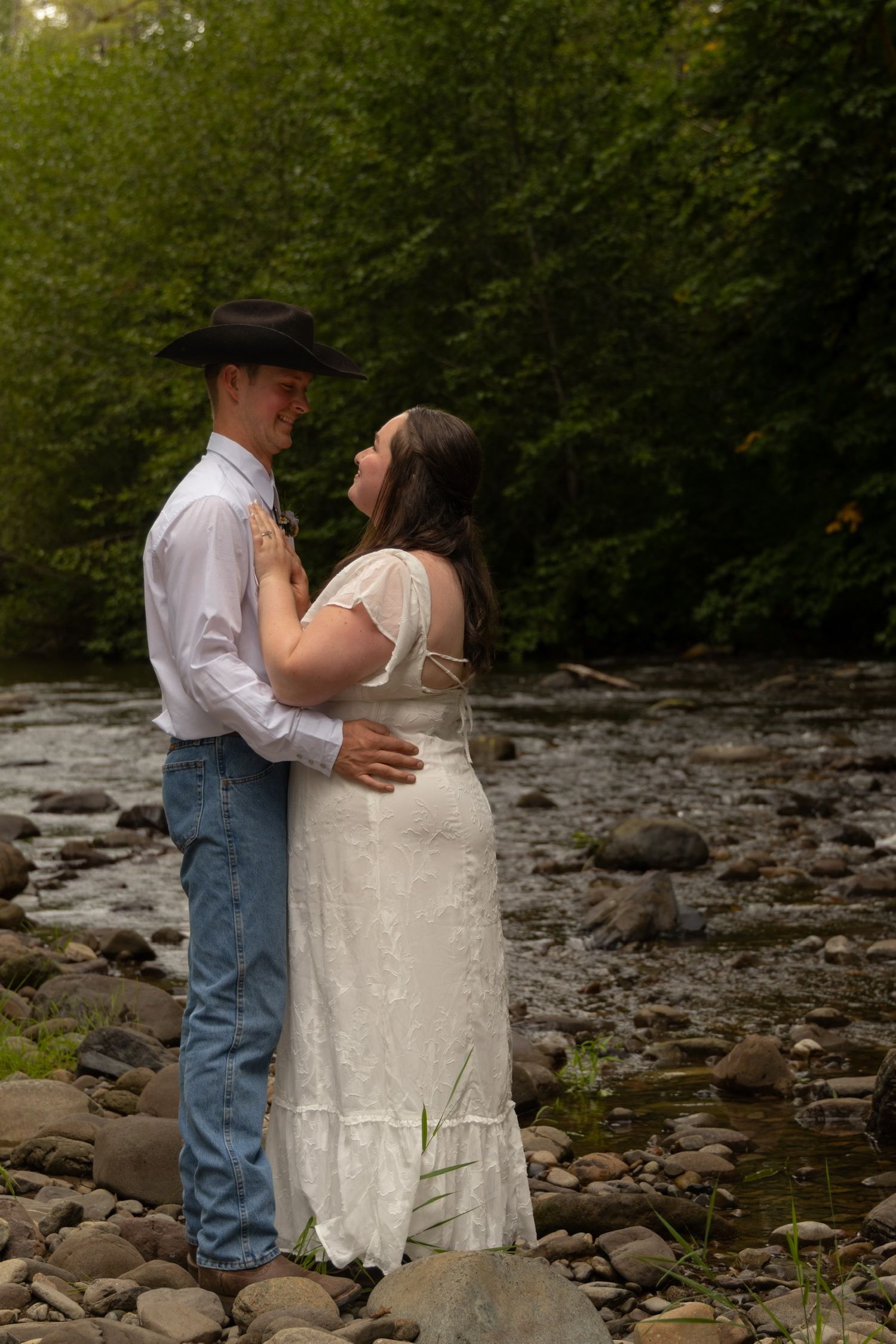 A couple embraces by a rocky riverbank in the woods, with the bride in white and groom wearing a cowboy hat and jeans.