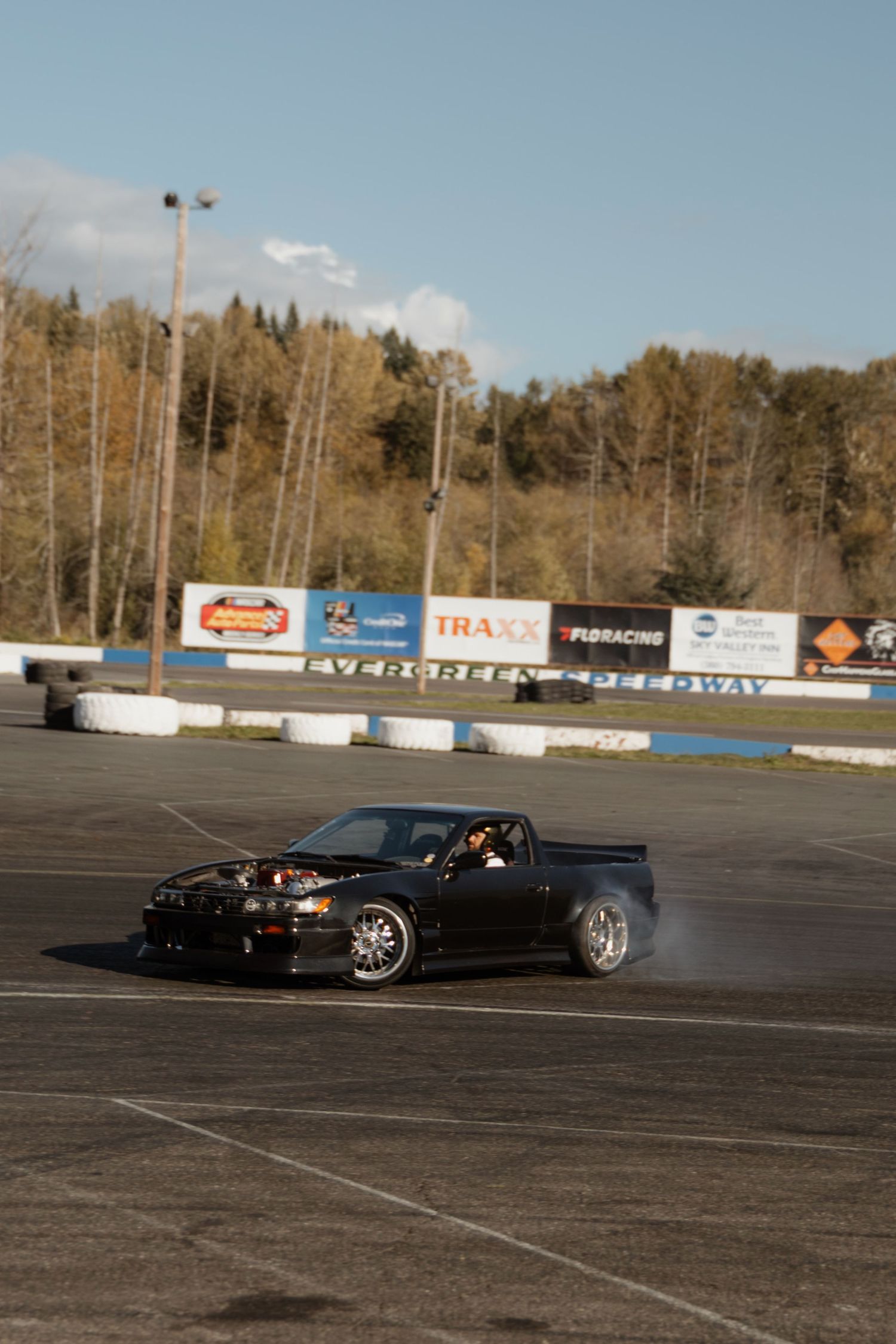 Black sports car drifting on a large empty asphalt lot with advertising banners and trees in the background.