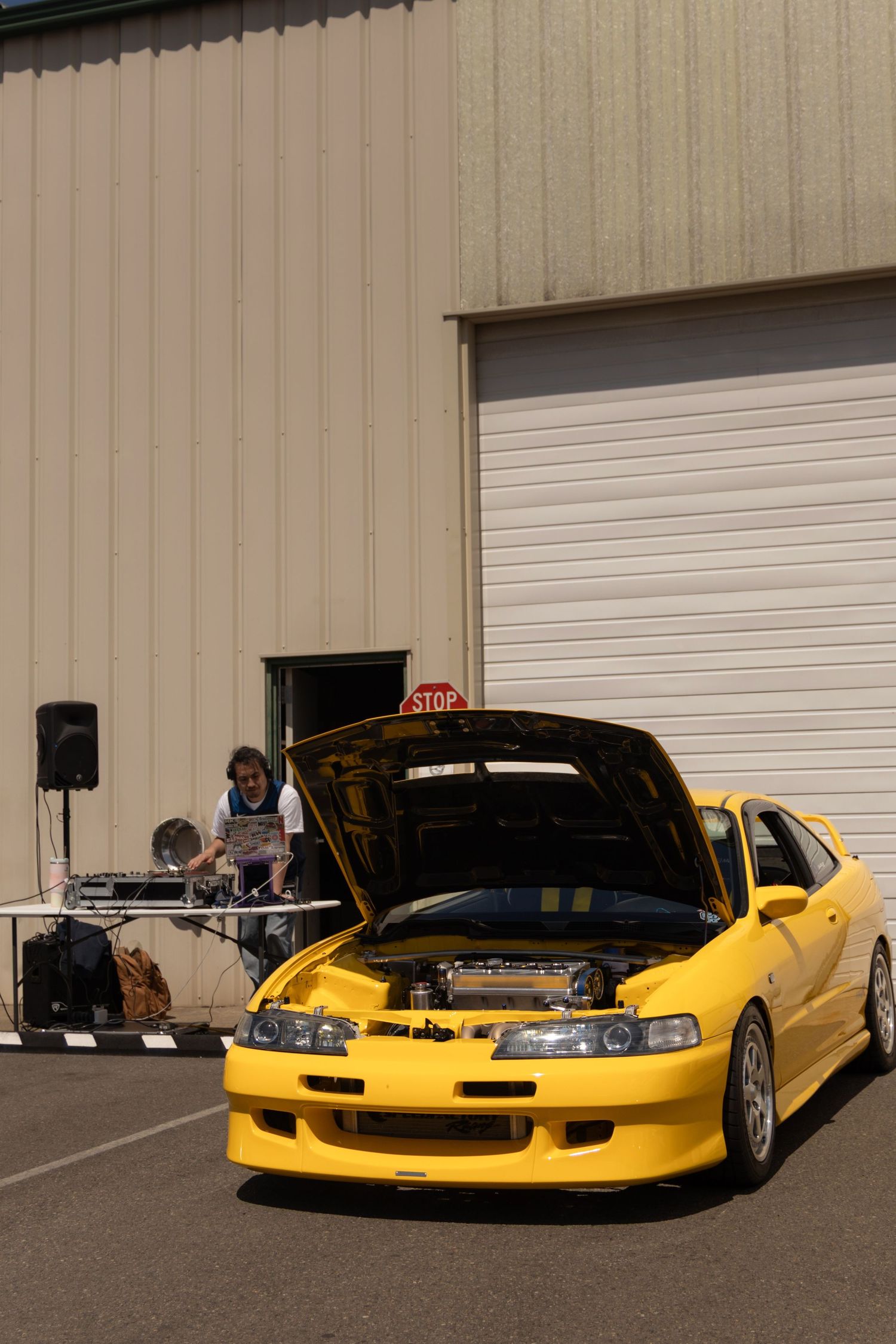 A bright yellow modified Honda Integra with an open hood parked in front of a garage door with DJ equipment visible.