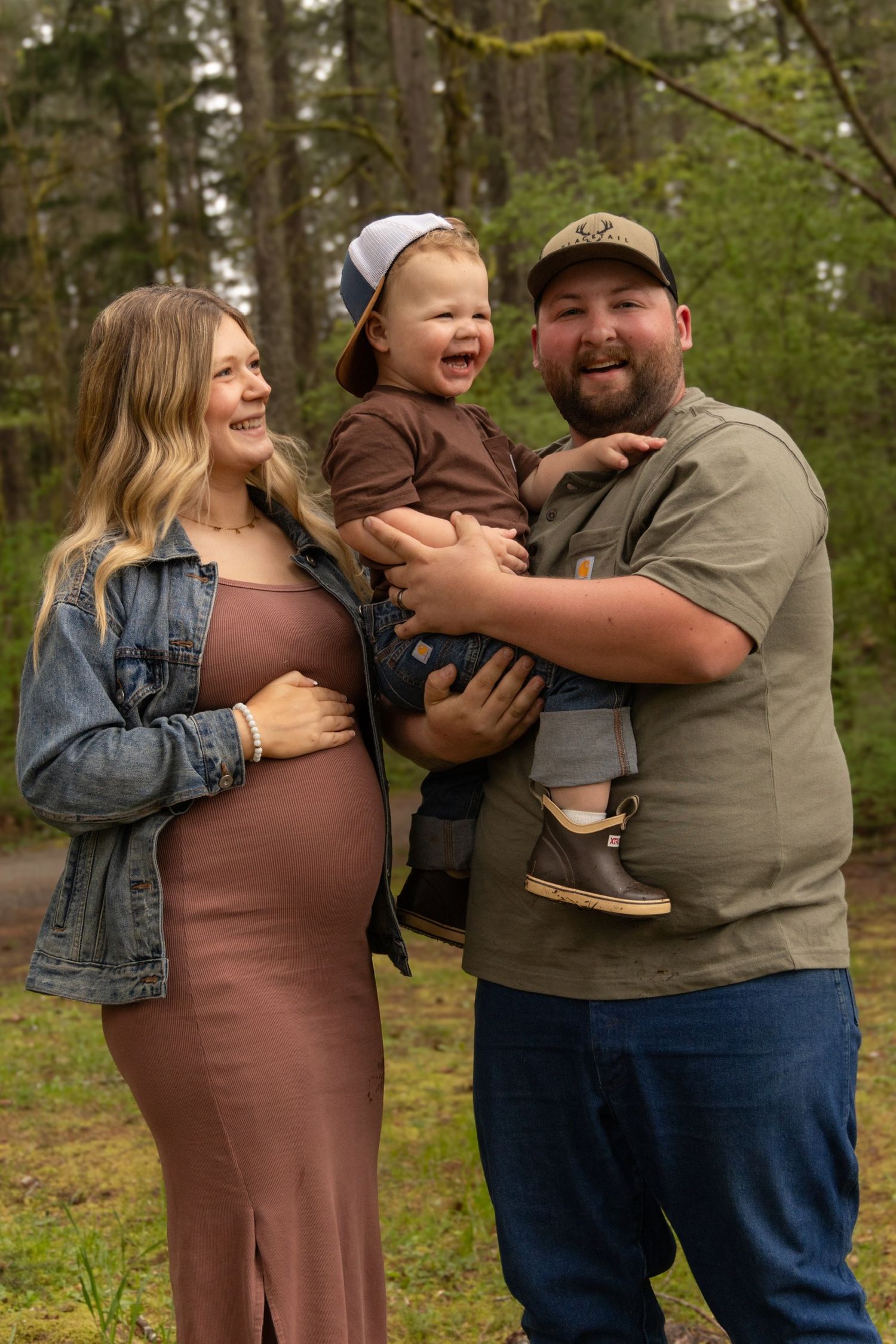 A joyful outdoor family moment in the woods with a pregnant woman in a brown dress and denim jacket.