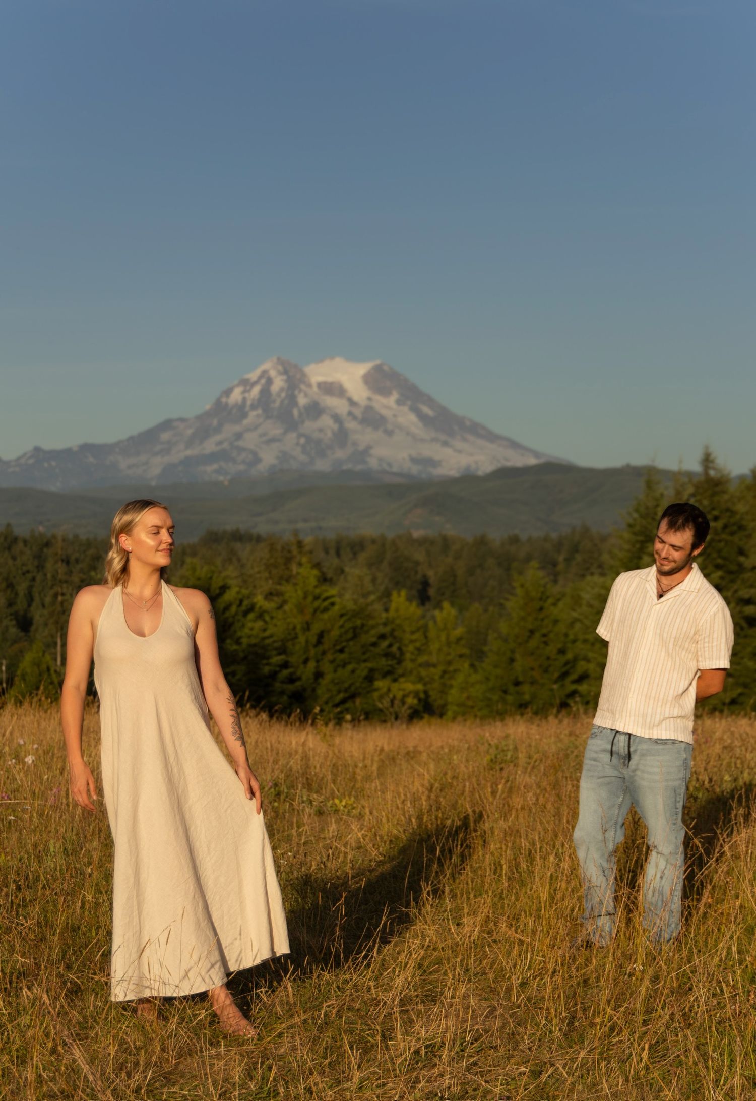 A couple in light summer clothing standing apart in a golden meadow with Mount Hood in the background.