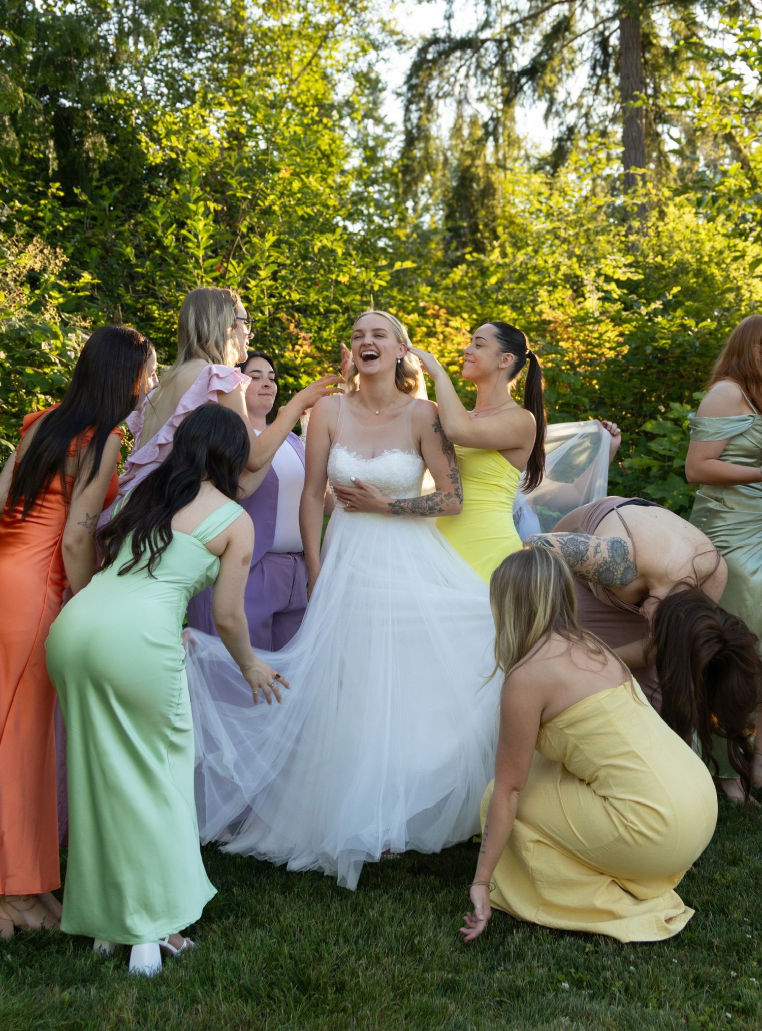 A group in colorful pastel bridesmaid dresses gather joyfully around a bride in a white gown during an outdoor celebration.