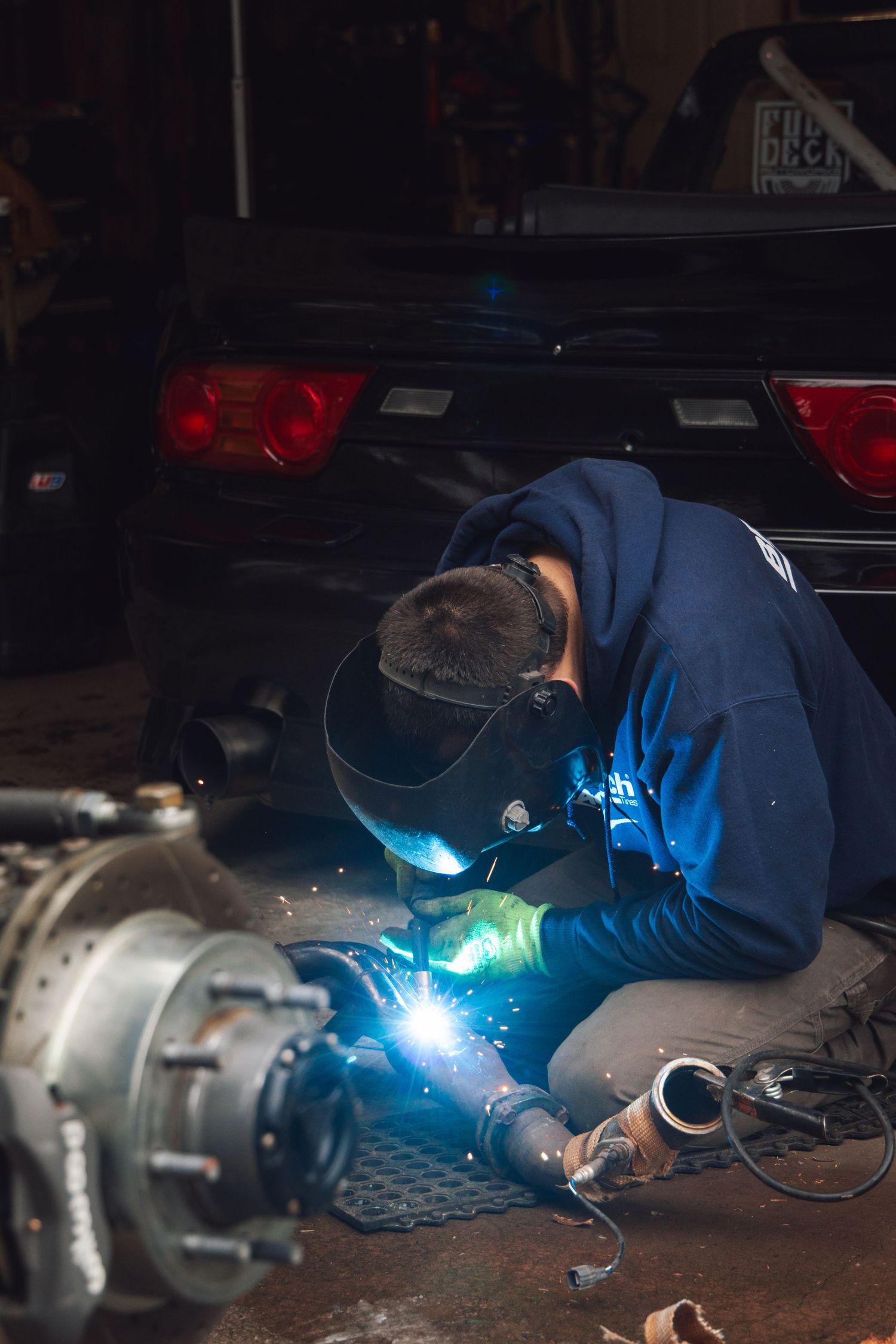 Welder working on car parts in a dimly lit garage with bright blue welding sparks.