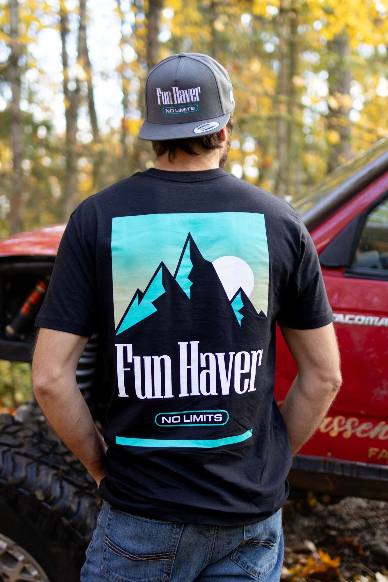 Black t-shirts with mountain logo designs shown outdoors against autumn foliage and trucks.