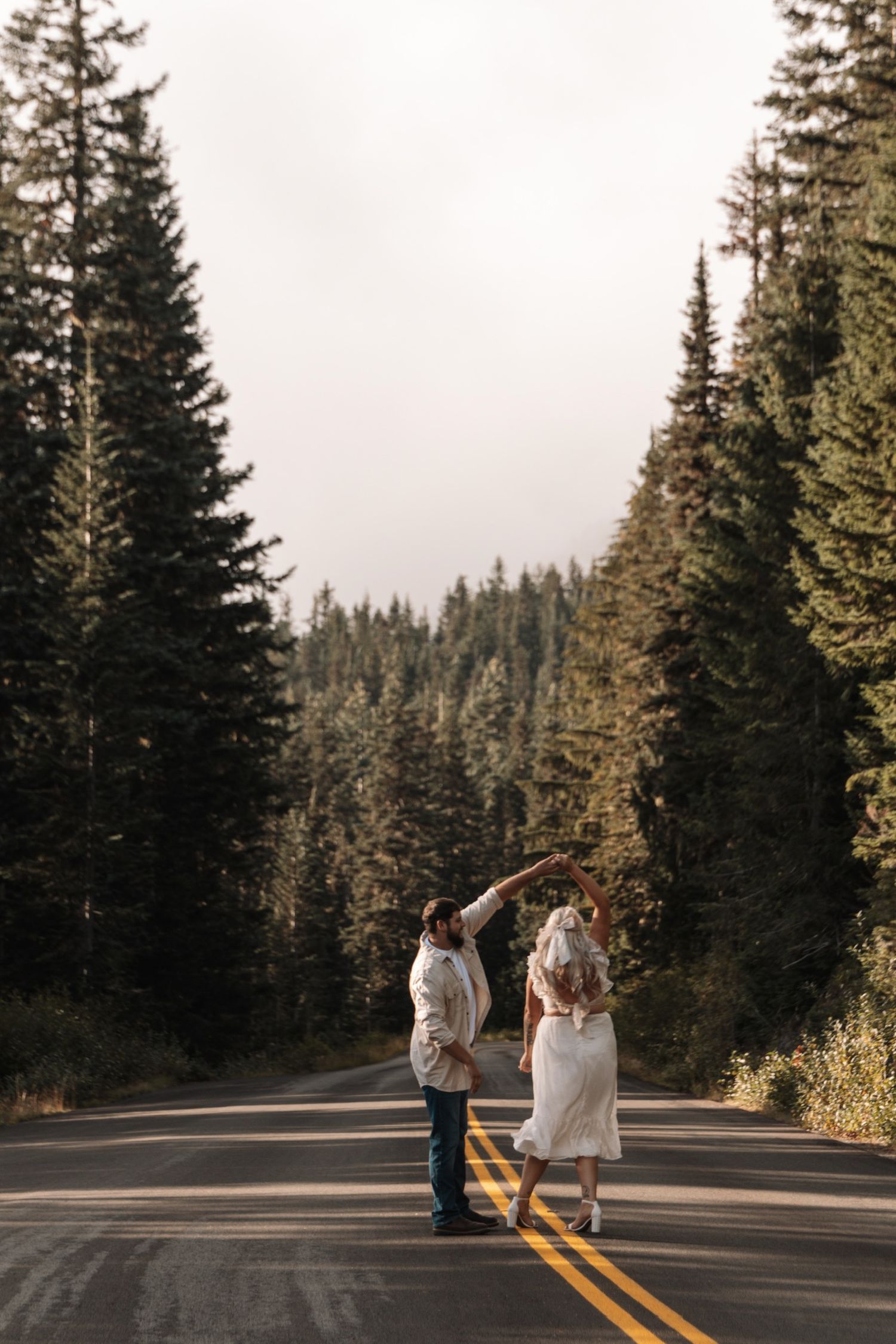 People walk down a scenic mountain road lined with tall evergreen trees on a misty day.