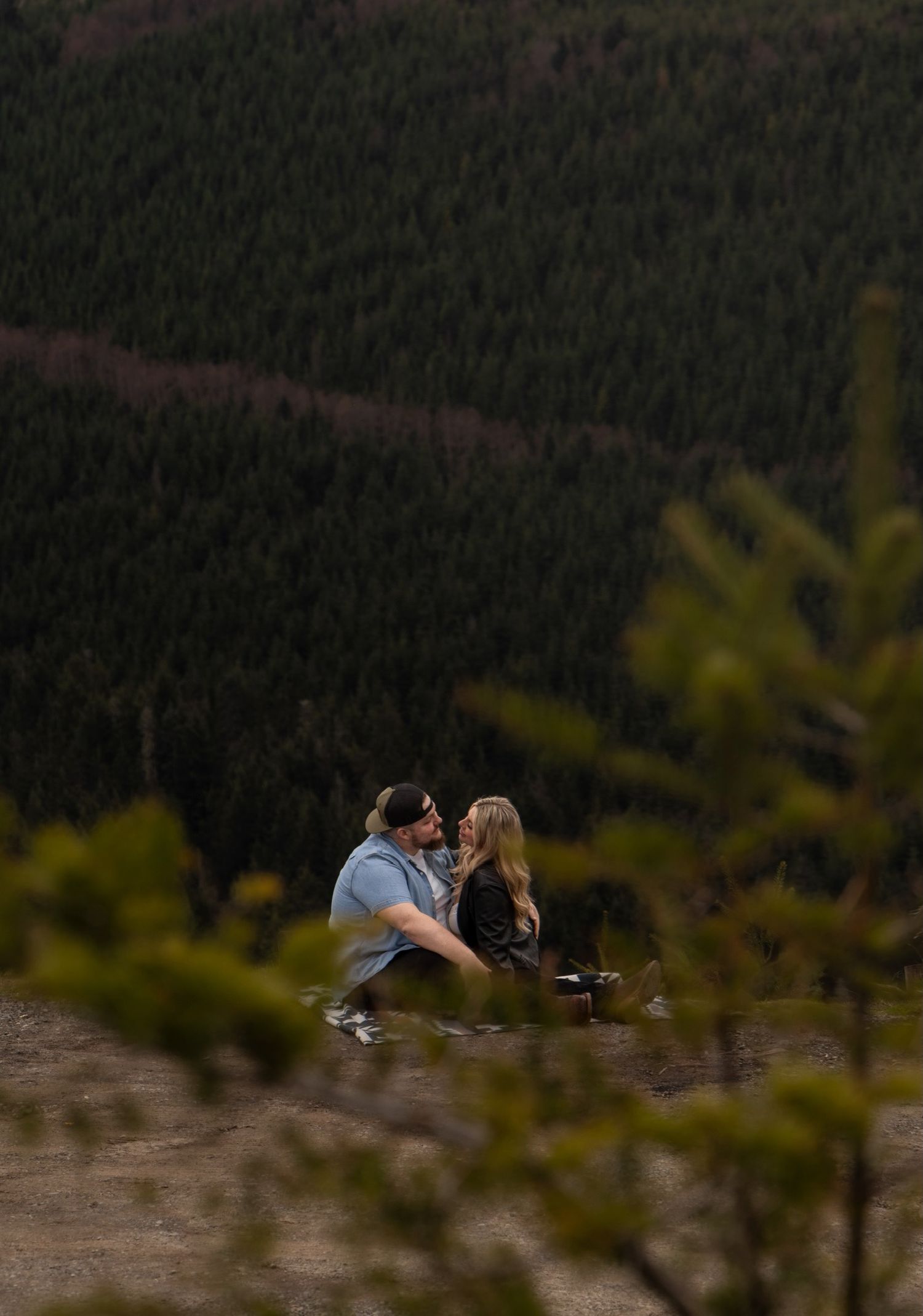 A couple shares a romantic moment on a hillside overlooking a dense forest landscape at sunset.