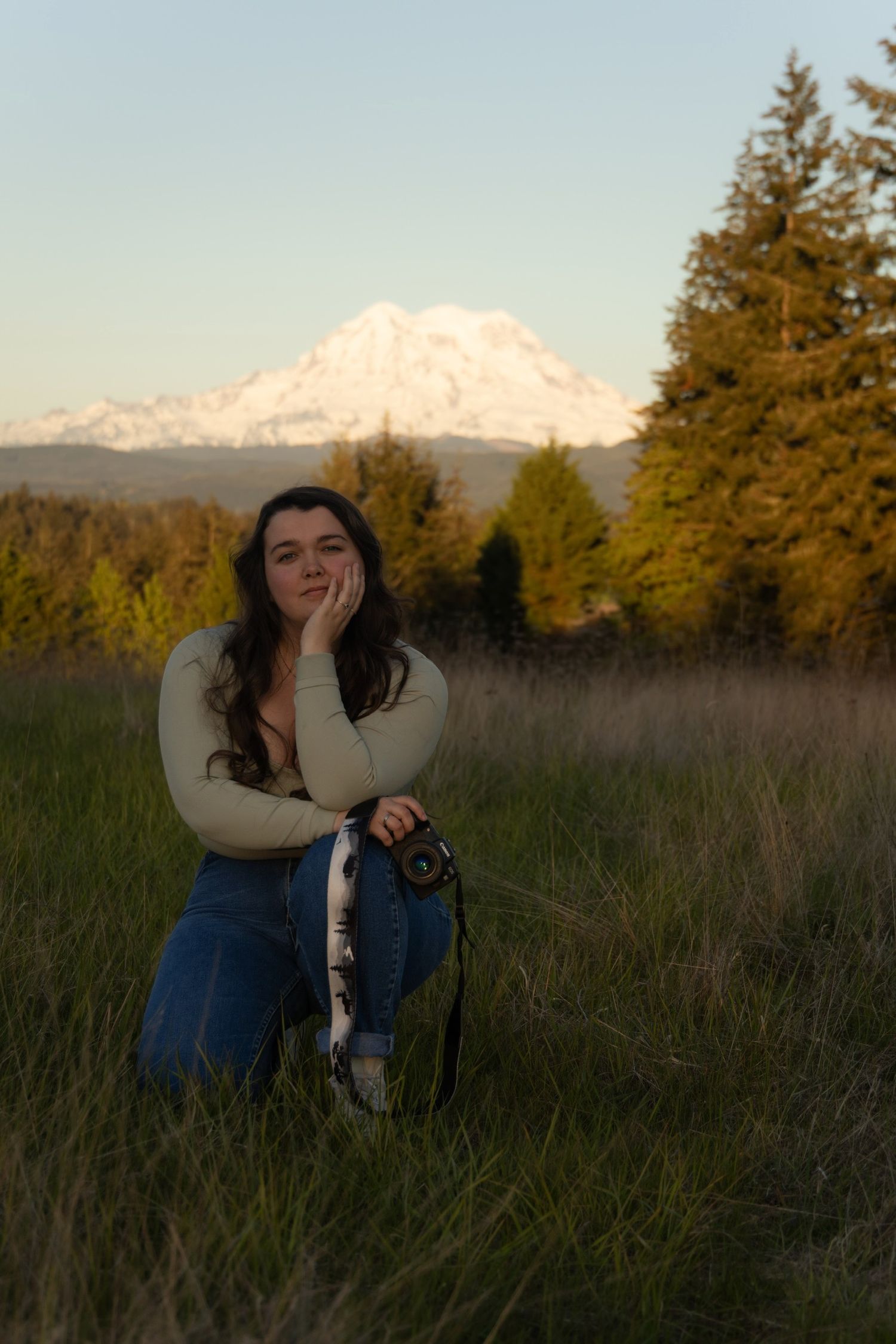 Person in cream sweater and jeans poses in field at sunset with mountain range visible in background.