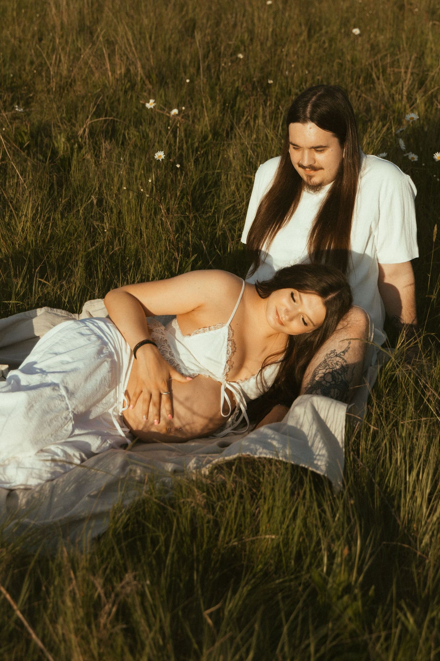 Couple relaxing in a sunny meadow, pregnant woman lying on blanket in white dress while partner sits beside her.