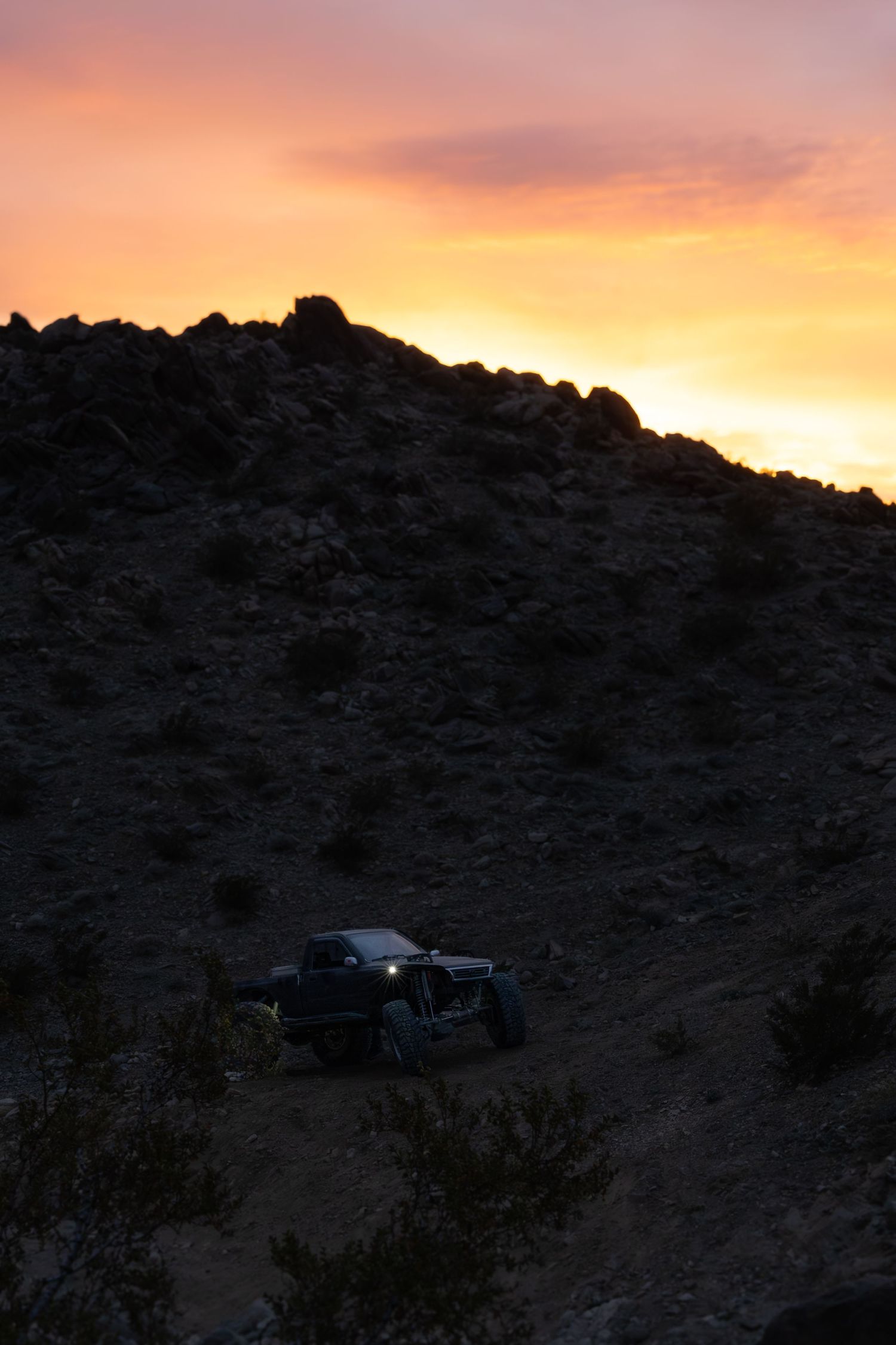 A dramatic desert sunset silhouettes rocky hills with vehicles parked along the ridge against an orange and pink sky.