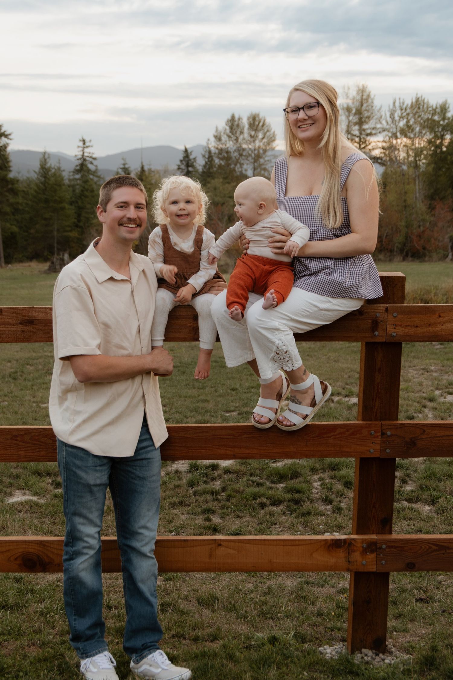A family shares joyful moments together while sitting and playing on a wooden fence in a rural field at sunset.