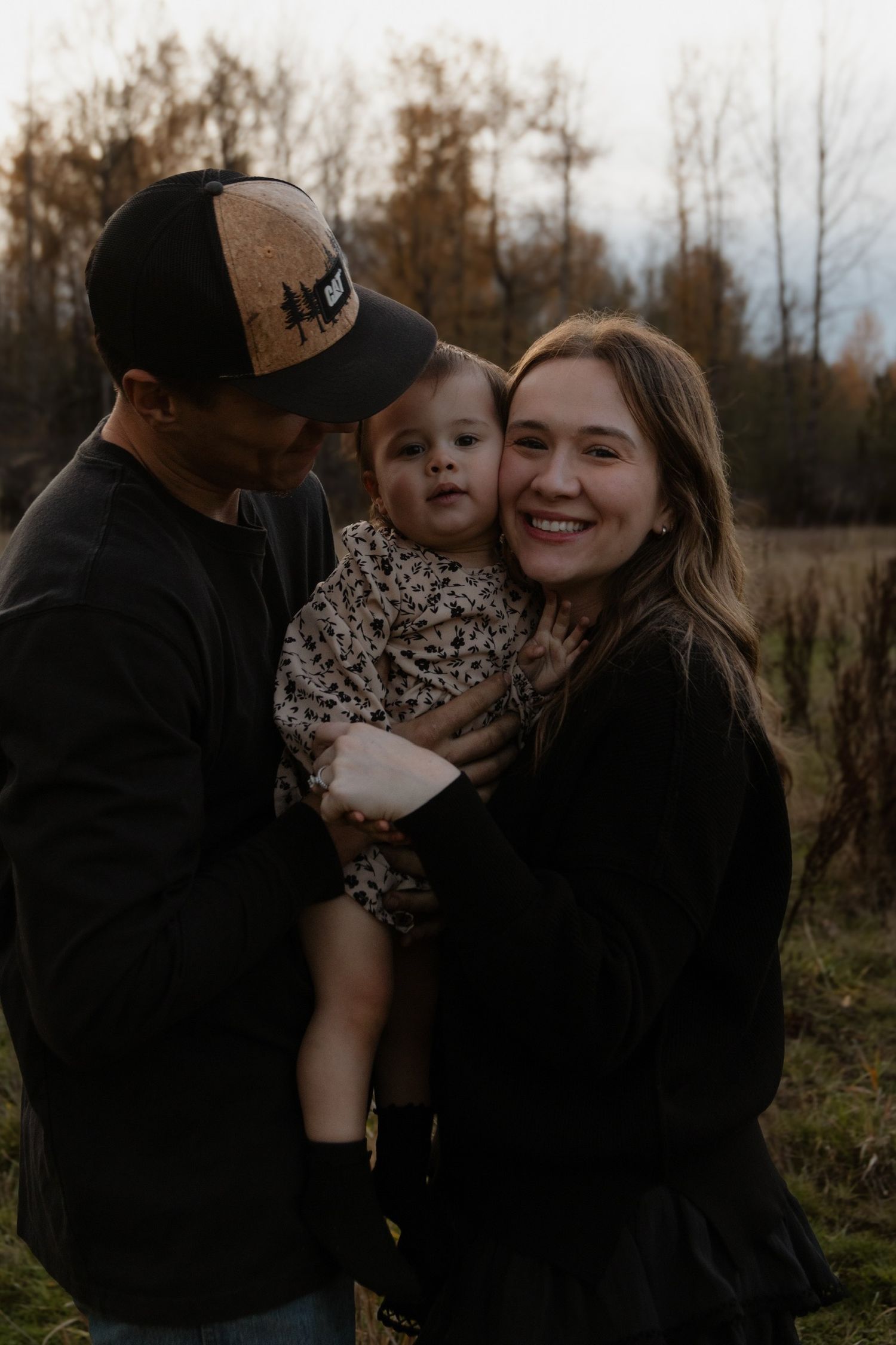 A family shares tender moments together in an outdoor setting with autumn foliage in the background.