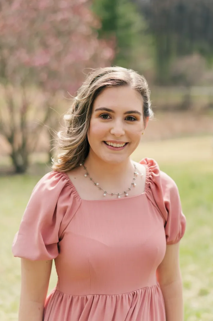 Young woman in a pink dress with a pearl necklace smiling outdoors with blooming trees in the background.
