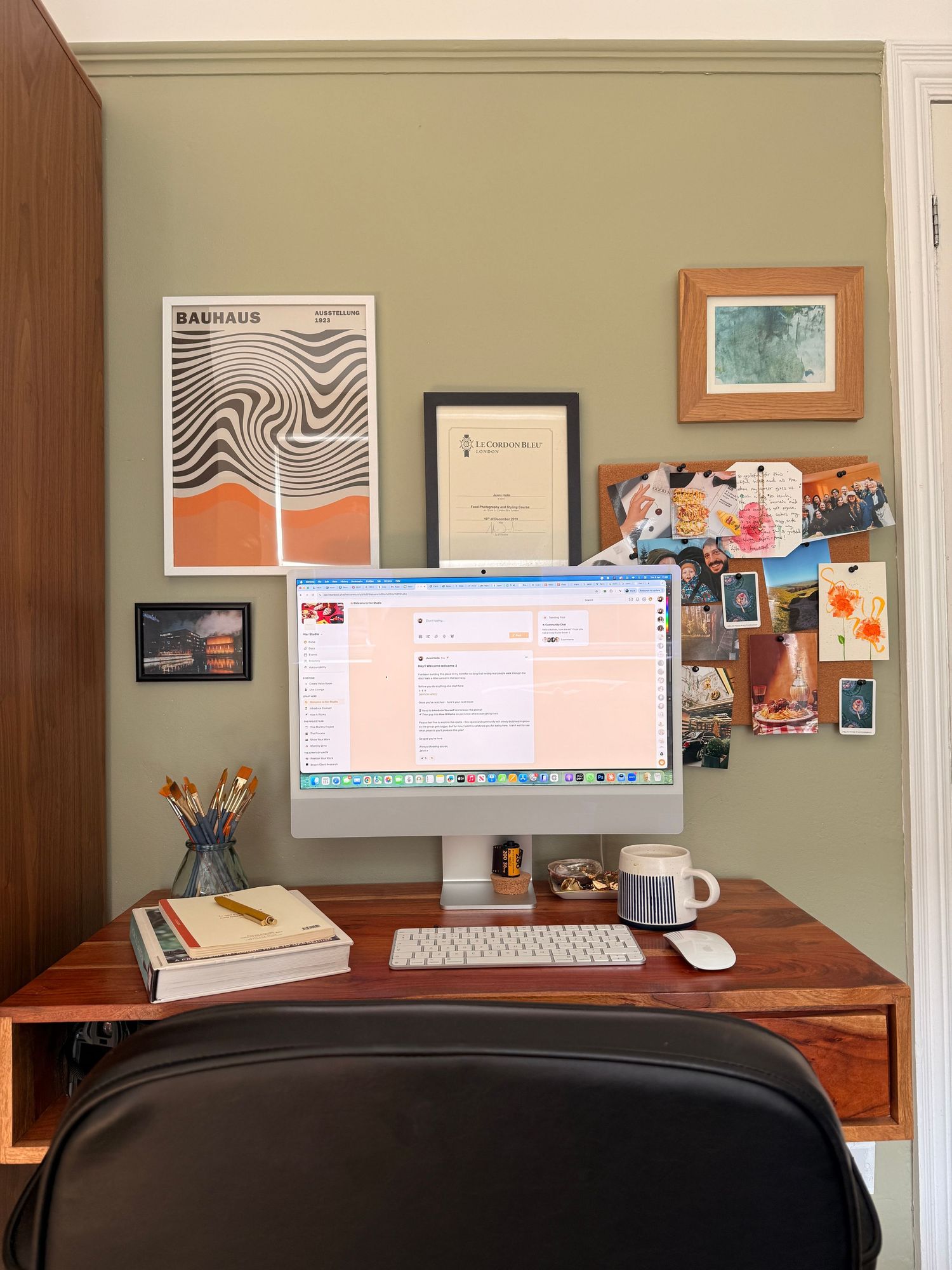 A cozy home office desk with an iMac, keyboard, and mouse, surrounded by art prints and a mood board on sage green walls.