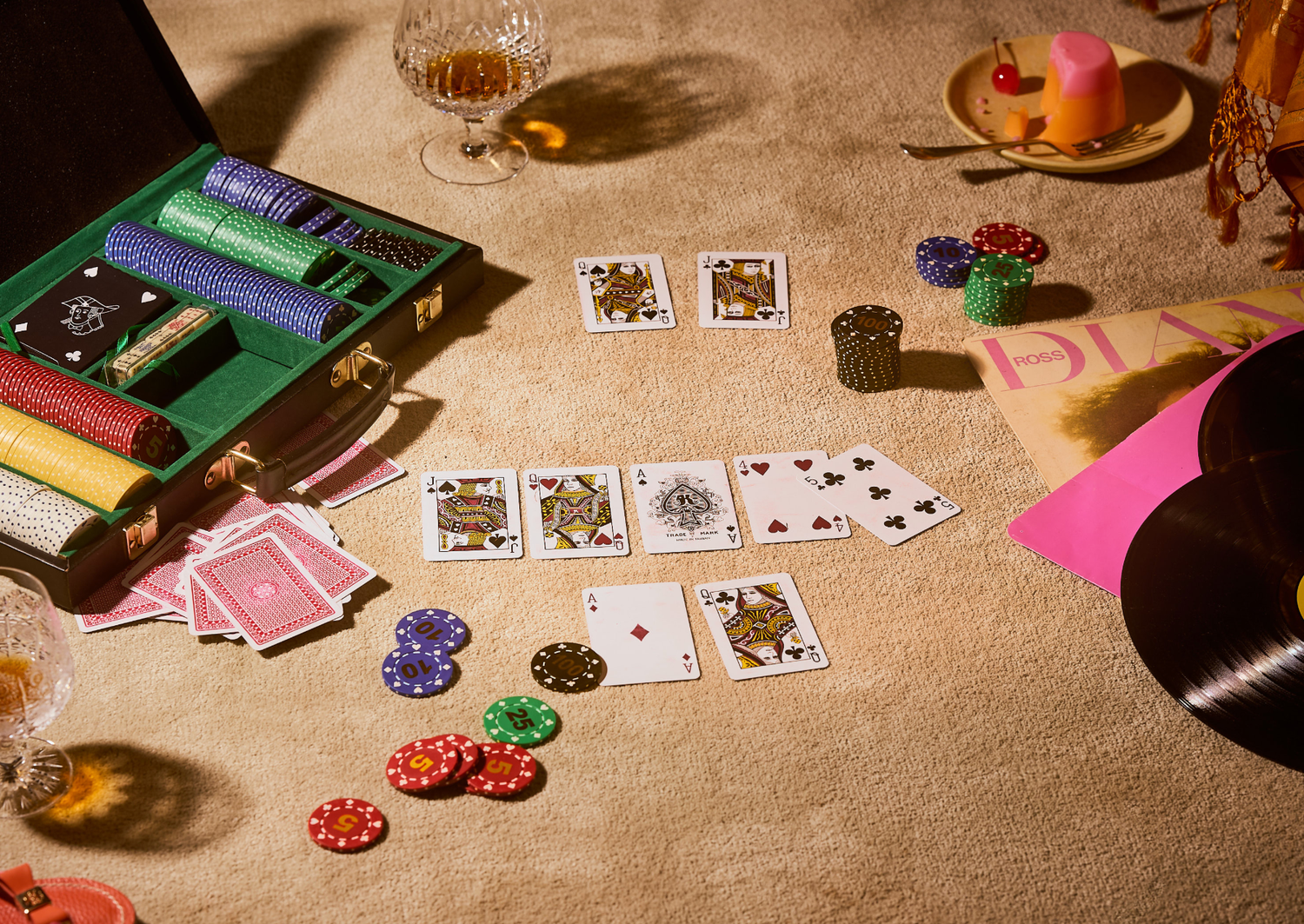 Poker chips and playing cards spread on a beige surface with a whiskey glass and pink decor nearby.