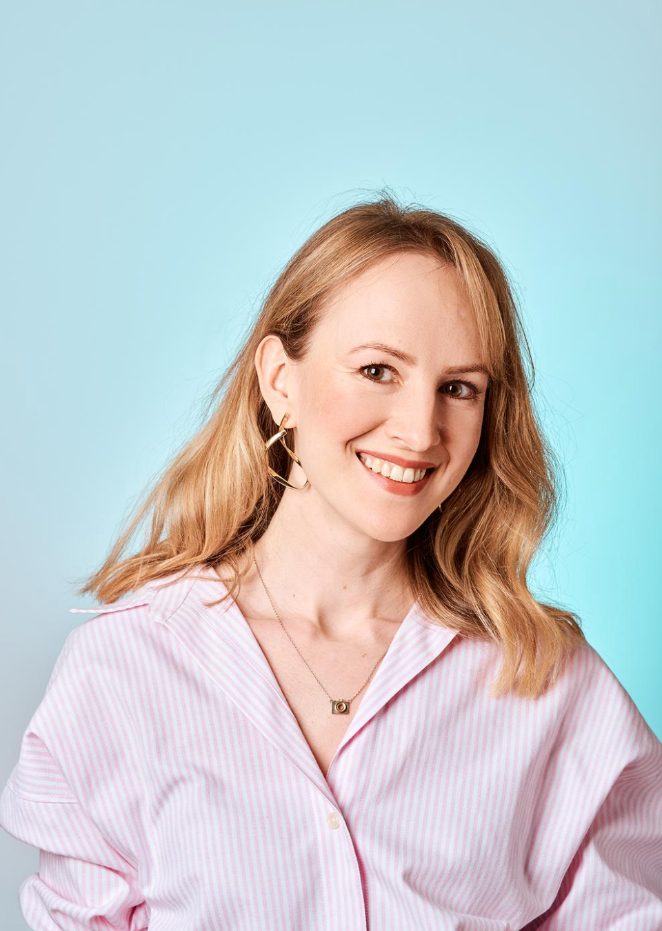 Smiling blonde woman in a pink striped shirt posing against a light blue background wearing gold hoop earrings.
