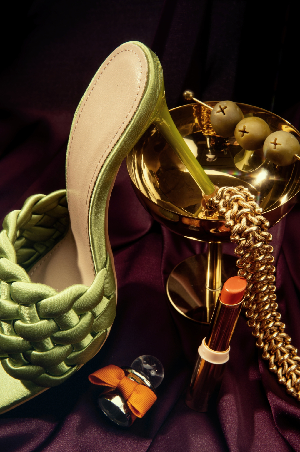 Green woven heeled mule shoe styled with gold chain necklace, orange lipstick, and decorative gold bowl on purple fabric.