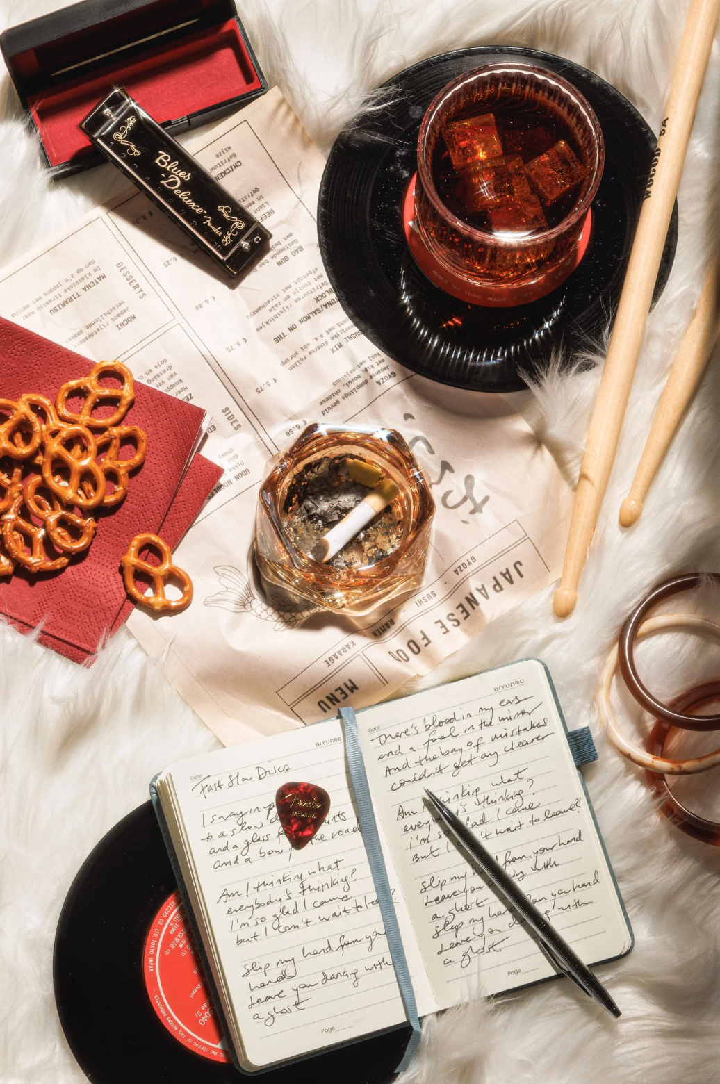 Flat lay of a journal, vinyl record, pretzels, drumsticks, drinks, and an ashtray on white fur.