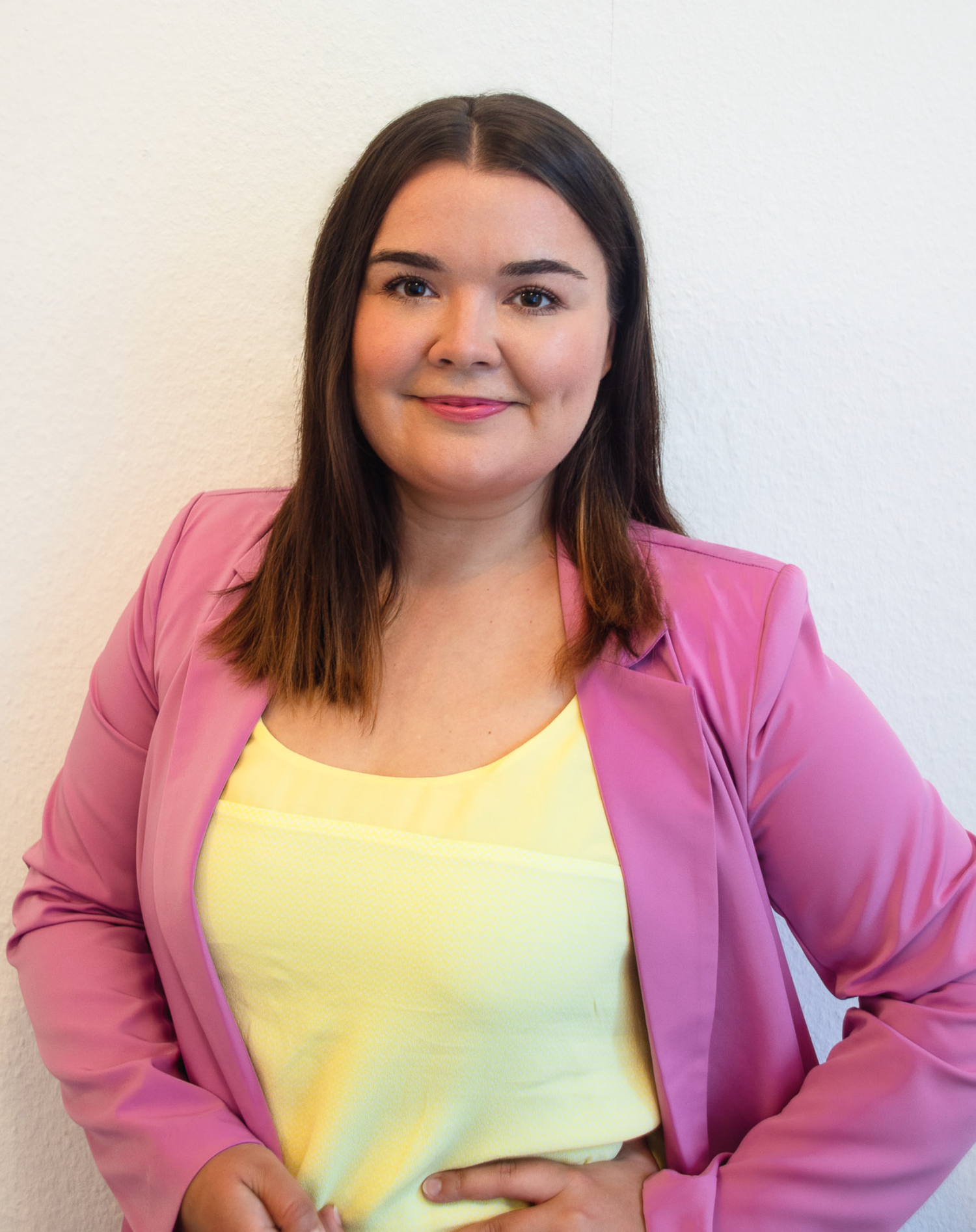 Young woman in a pink blazer and yellow top smiling confidently while standing against a white wall.
