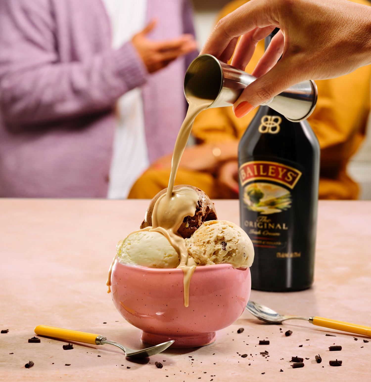 Baileys Original Irish Cream being poured over a pink bowl of ice cream scoops with chocolate pieces on a pink surface.